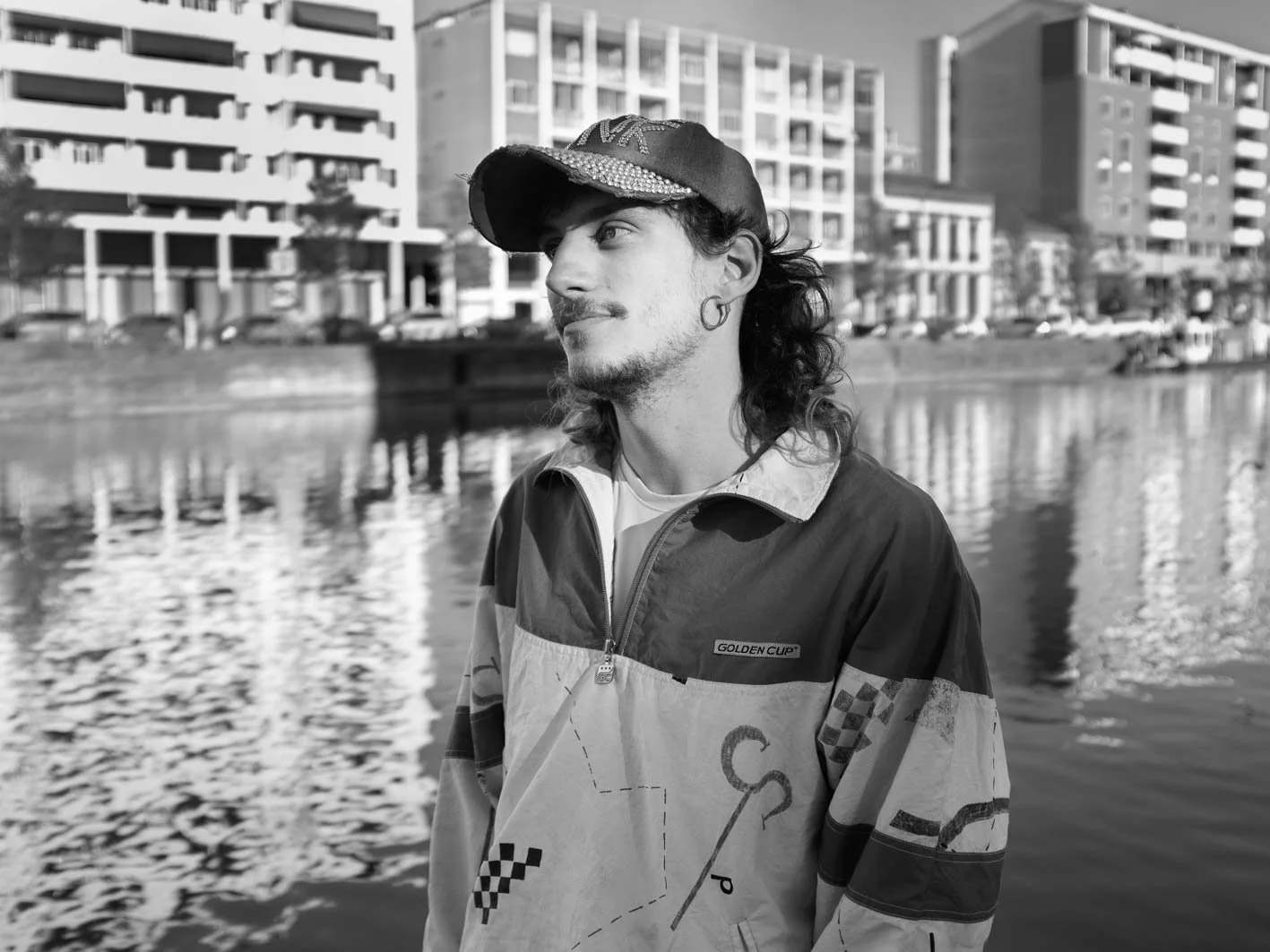 Un homme avec chapeau et boucle d'oreille, debout près d'une rivière, avec des bâtiments modernes en arrière-plan.