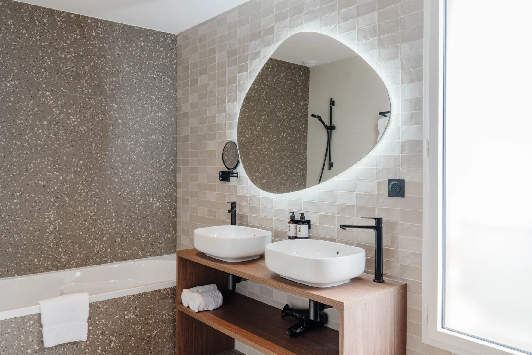 Salle de bain moderne avec deux lavabos ovales en céramique sur une meuble en bois, miroir rond avec éclairage intégré, douche avec paroi en verre, mur en carreaux beige et mur à carreaux terrazzo, prises électriques noires, serviettes blanches.