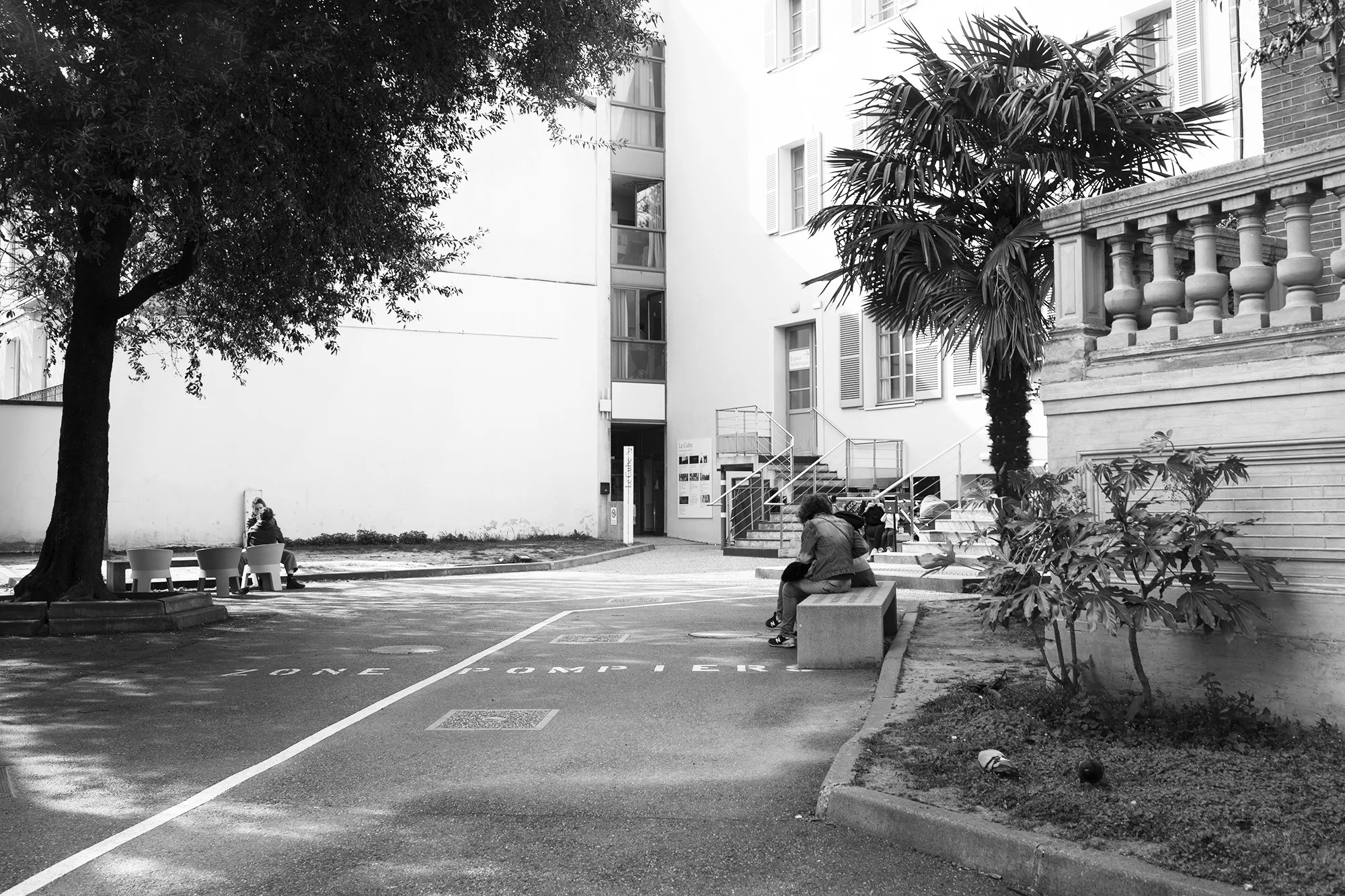 Une cour intérieure avec des personnes assises sur des bancs, entourée d'arbres et de bâtiments résidentiels. L'image est en noir et blanc.