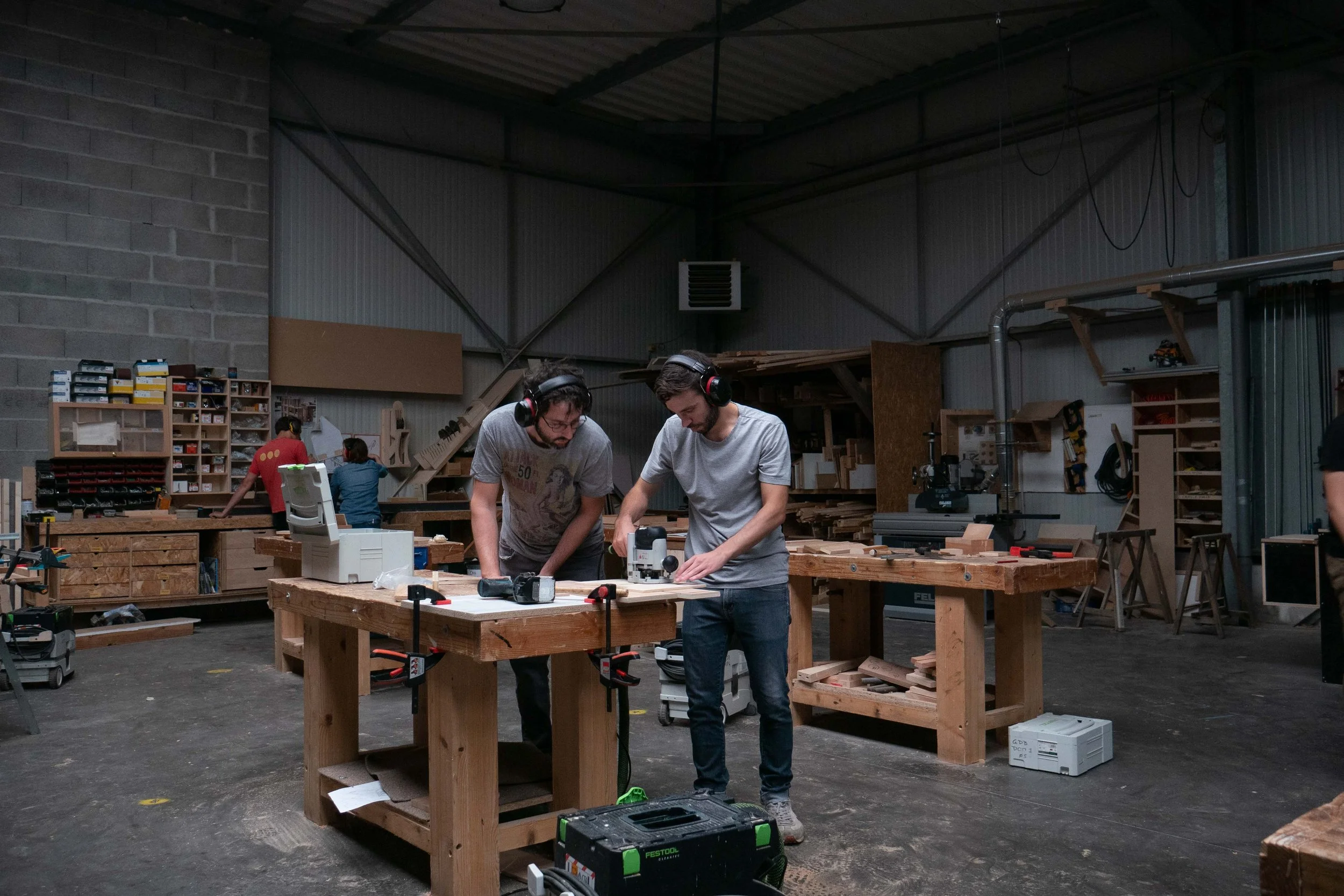Deux jeunes hommes travaillent à une table en bois dans un atelier de menuiserie avec divers outils et matériaux autour, portant des casques de protection auditifs.