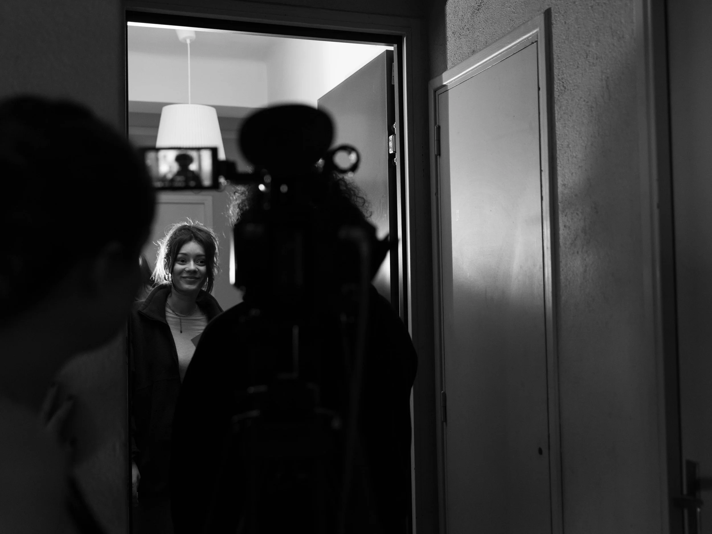 Une femme souriante regarde à travers une caméra située dans un couloir. La photo est en noir et blanc, avec un décor intérieur simple.