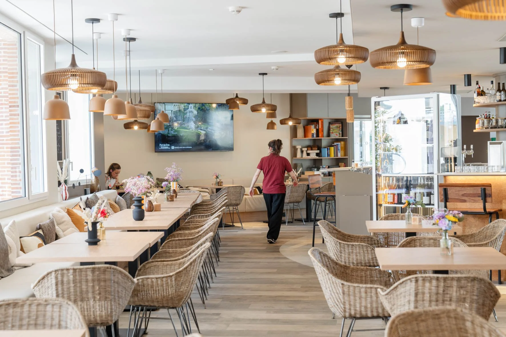 Intérieur d'un restaurant ou café avec tables, chaises en rotin, décor floral, lampes suspendues en bois, écran plat au mur, et personnel en uniforme