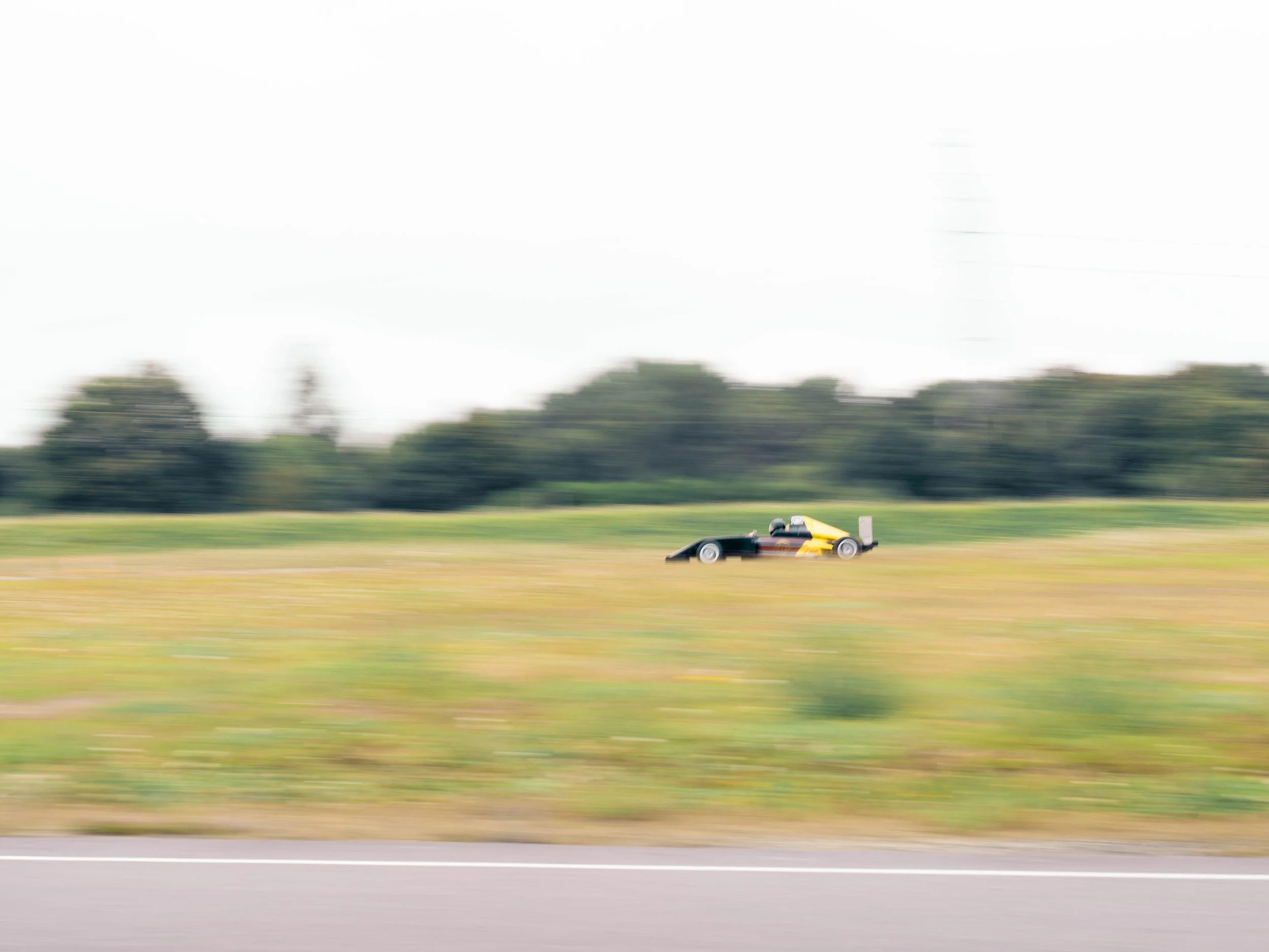 Voiture de course en mouvement sur une route, arrière-plan flou avec des arbres et un ciel nuageux.
