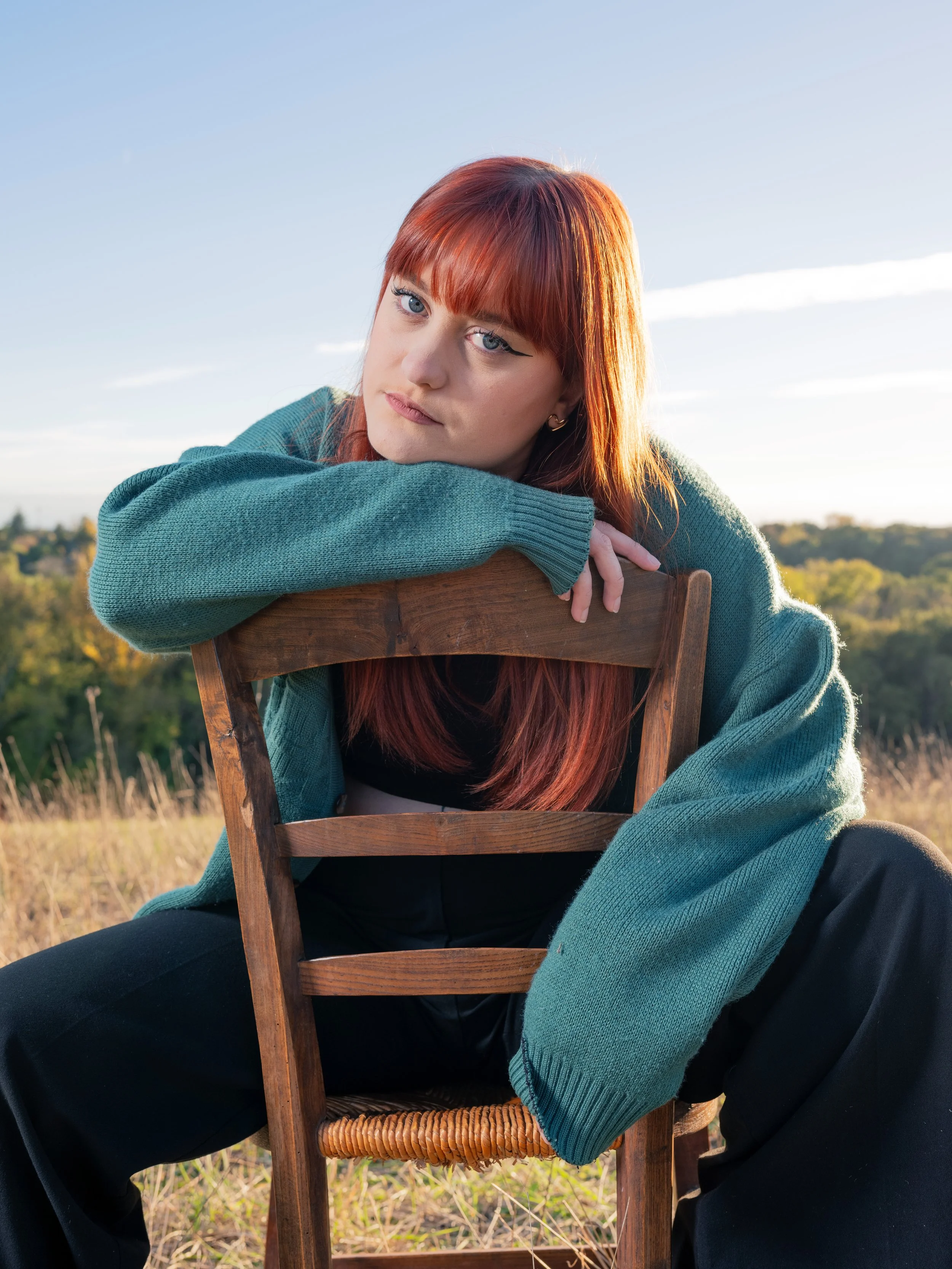 Femme aux cheveux roux, portant un pull vert, assise sur une chaise en bois dans un champ avec un ciel bleu en arrière-plan.