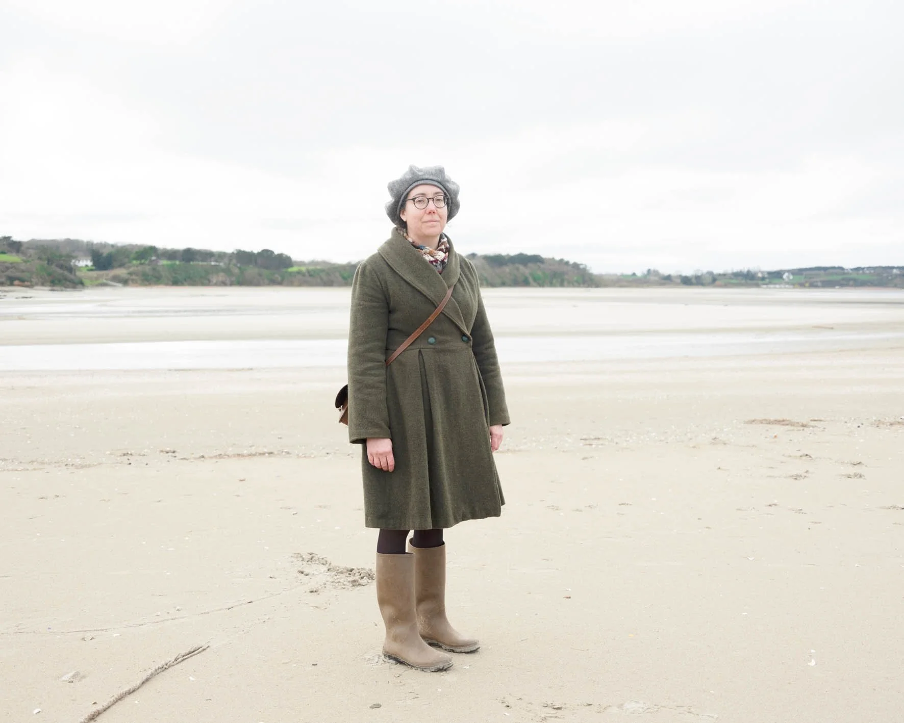 Une femme portant un manteau vert, des bottes en caoutchouc beige, une écharpe, un béret gris, et des lunettes, debout sur une plage déserte sous un ciel gris.