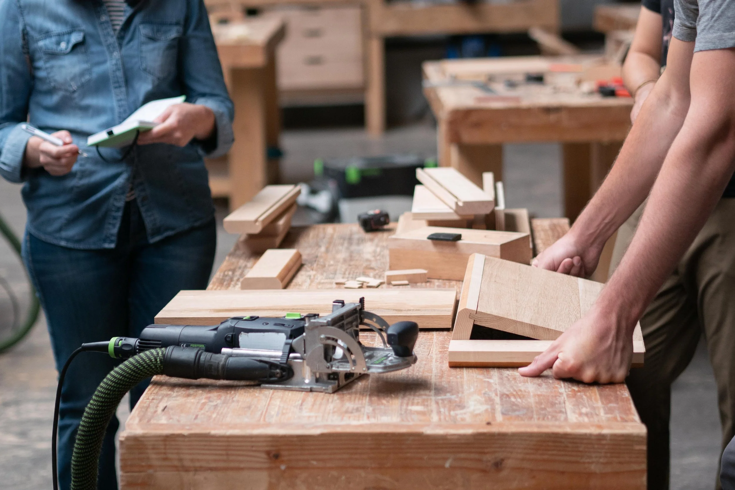 Personnes travaillant dans un atelier de menuiserie, utilisant des outils électriques et des blocs de bois sur une table.