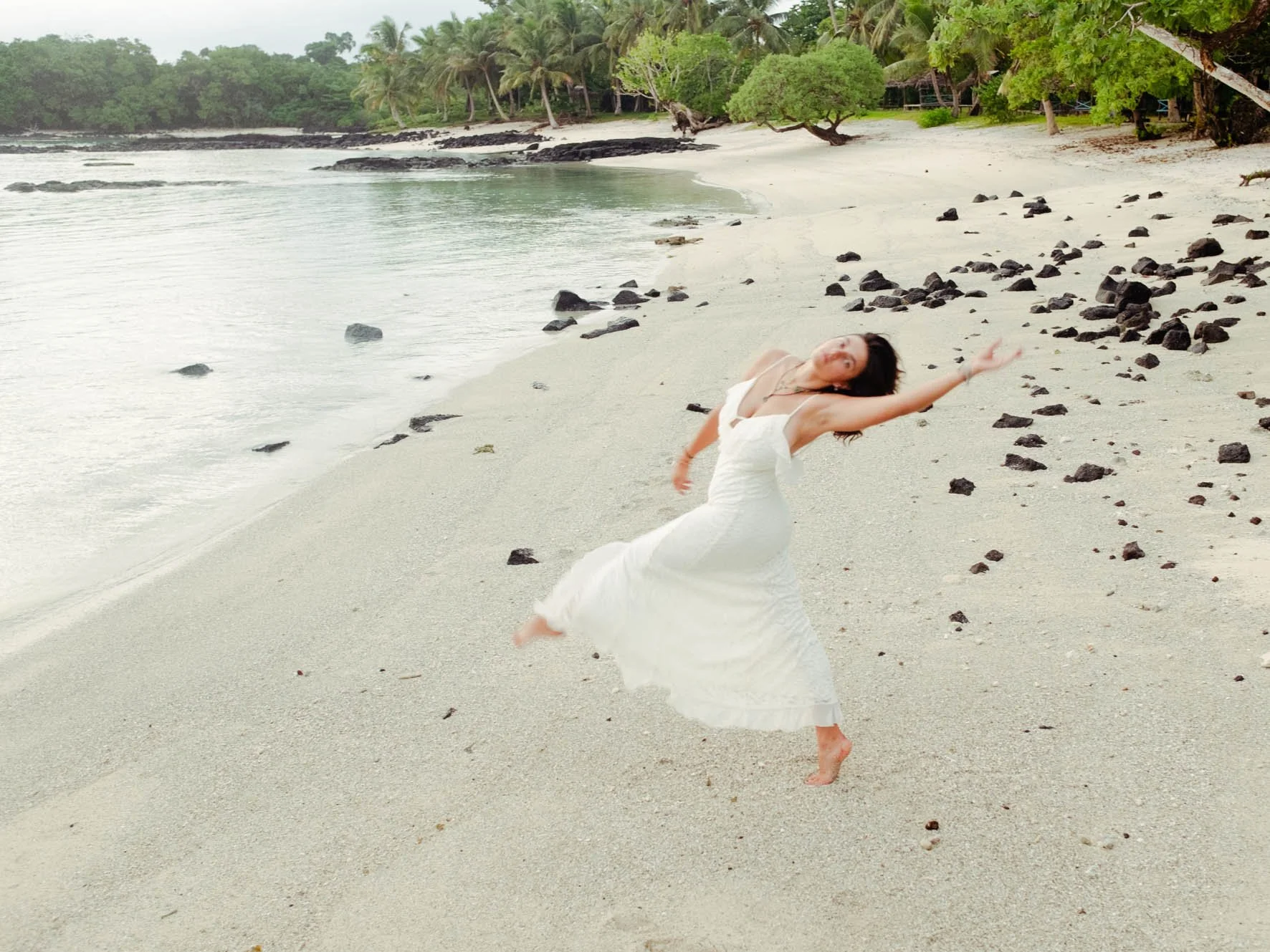 Une femme en robe blanche danse sur une plage de sable avec des rochers, des palmiers et une végétation verte en arrière-plan.