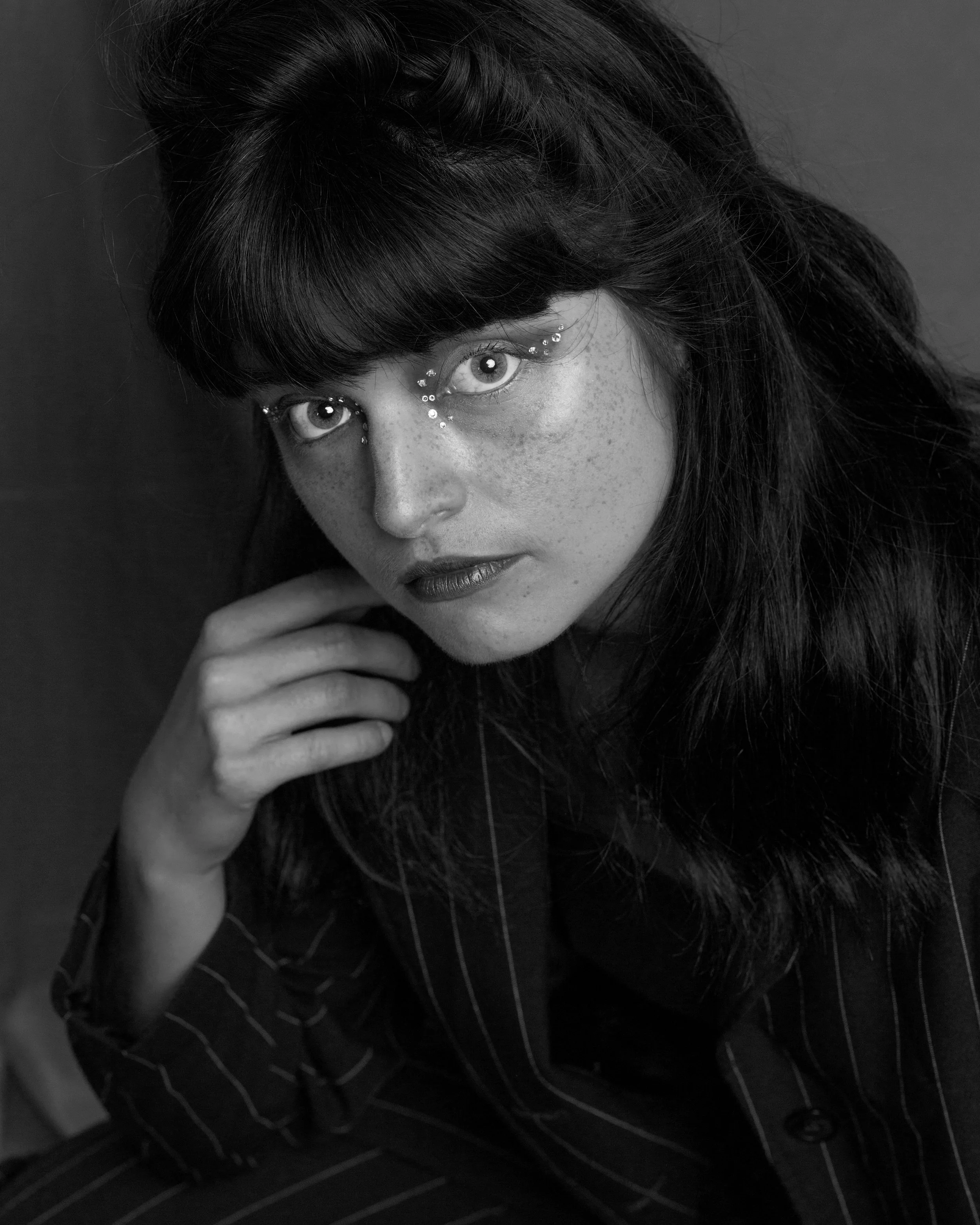 Un portrait en noir et blanc d'une femme avec de longs cheveux, portant des vêtements à rayures, regardant fixement la caméra, avec des détails de maquillage et de bijoux autour des yeux.