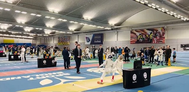 An indoor fencing competition taking place in a sports hall filled with spectators and participants, with banners and seating visible in the background.
