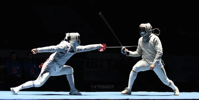 Two fencers in full protective gear sparring during a match, with one lunging forward and the other preparing to strike.
