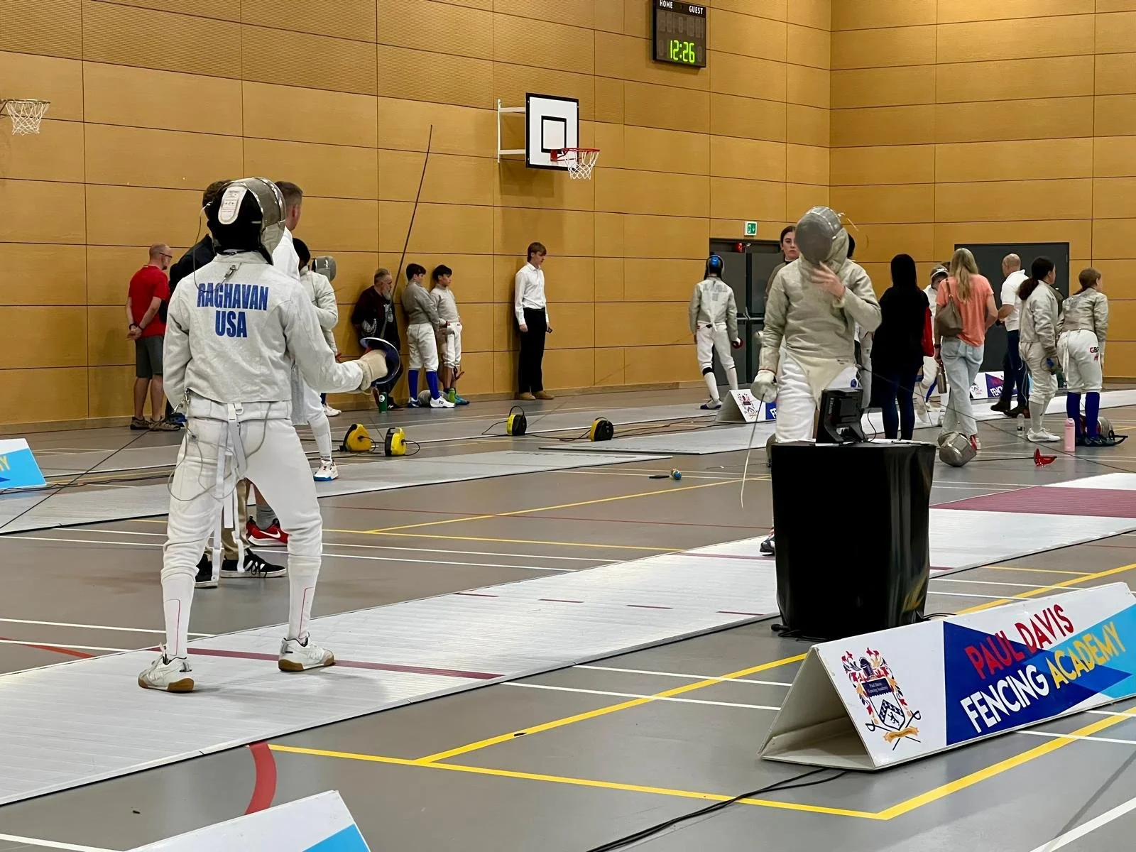 Fencers preparing for a match at an indoor fencing competition, with spectators and officials in the background, and a scoreboard showing 12:26.
