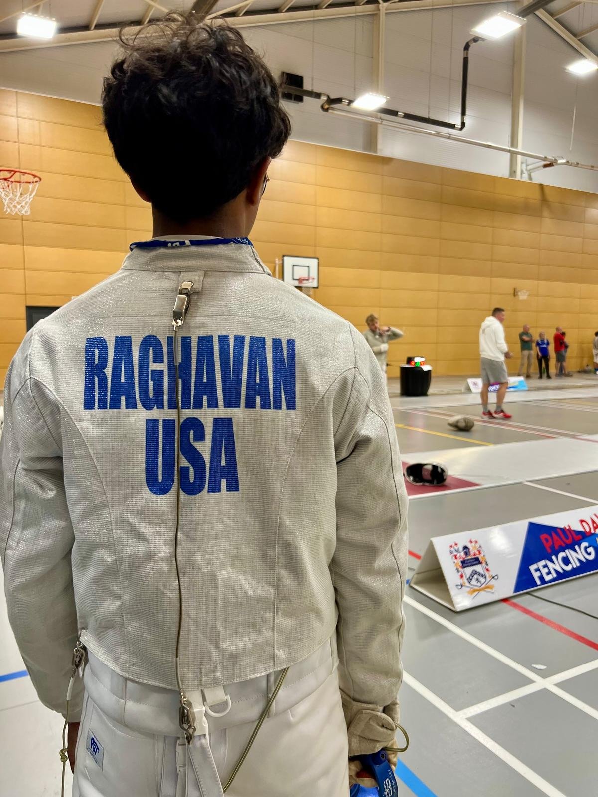 A fencer from the USA, Raghavan,, wearing a beige fencing jacket with his name and country on the back, standing in a sports hall during a fencing competition. Other people and fencing equipment are visible in the background.