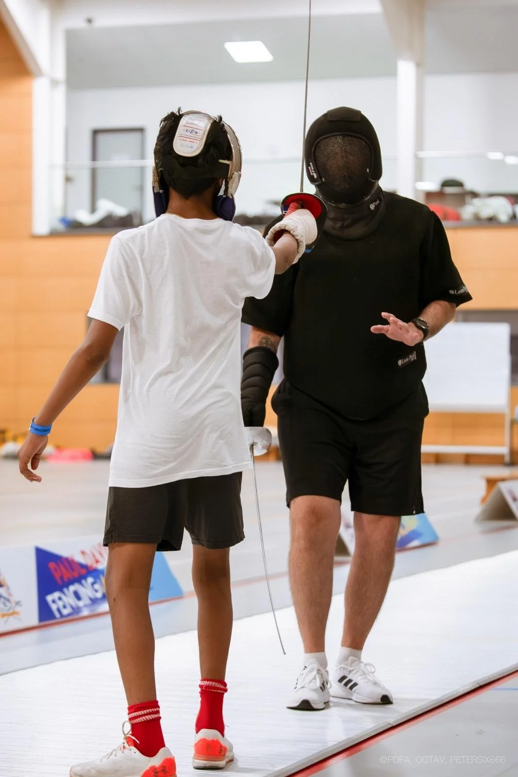 A young fencer in protective gear, including a mask and jacket, practicing with an instructor who is wearing fencing gear, on an indoor fencing strip.