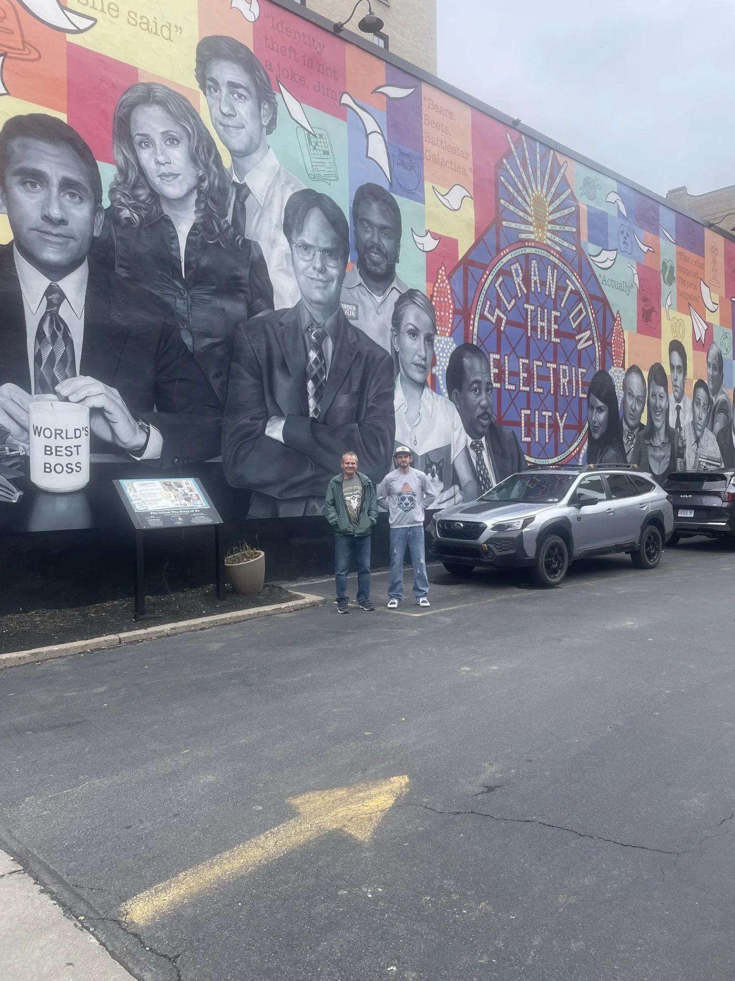 A large mural featuring black and white portraits of diverse people painted on a building wall. Two men are standing in front of the mural, with two cars parked nearby and a small potted plant beside the mural.