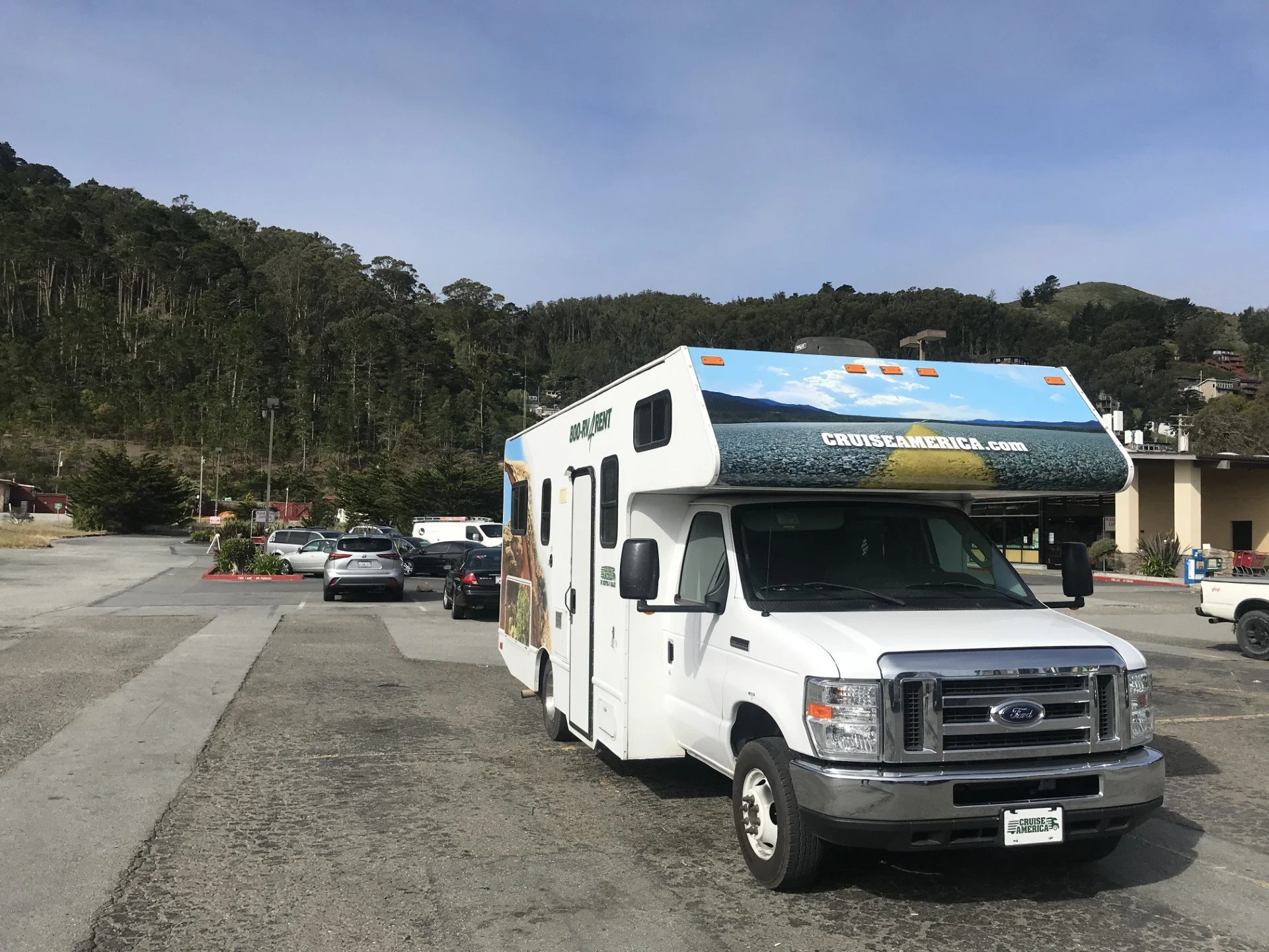 A parking lot with various cars and a white RV with a large scenic landscape mural on its side, parked under a clear sky with hills and trees in the background.