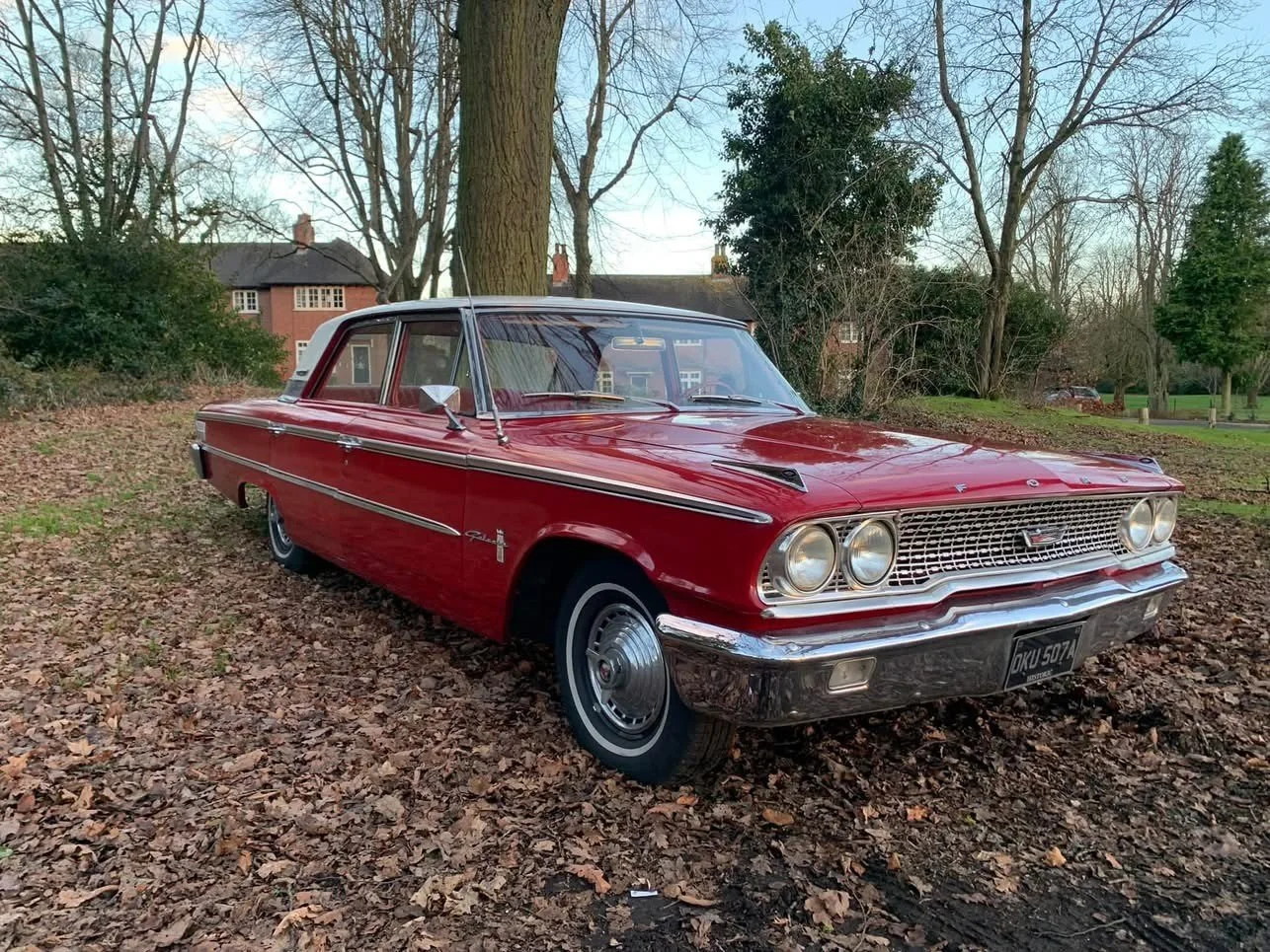 Red vintage Pontiac car parked on a ground covered with fallen leaves in a park-like setting with trees, grass, and residential houses in the background.