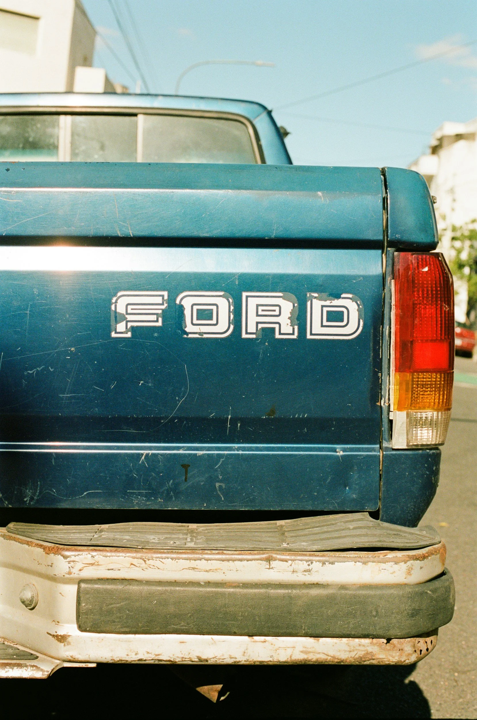 The back of a vintage blue Ford truck with visible scratches and a two-tone paint job, parked next to an old, white vehicle.