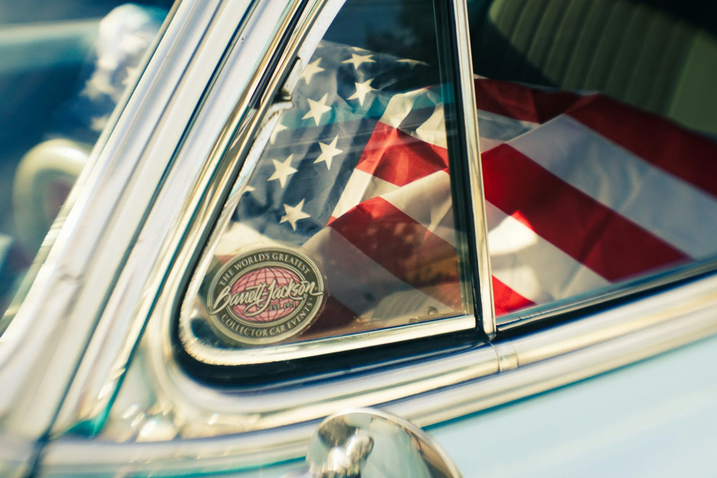 Close-up of a vintage car with an American flag in the background visible through the window. There is a sticker on the car window that reads 'The World's Greatest Collector Car Event'.