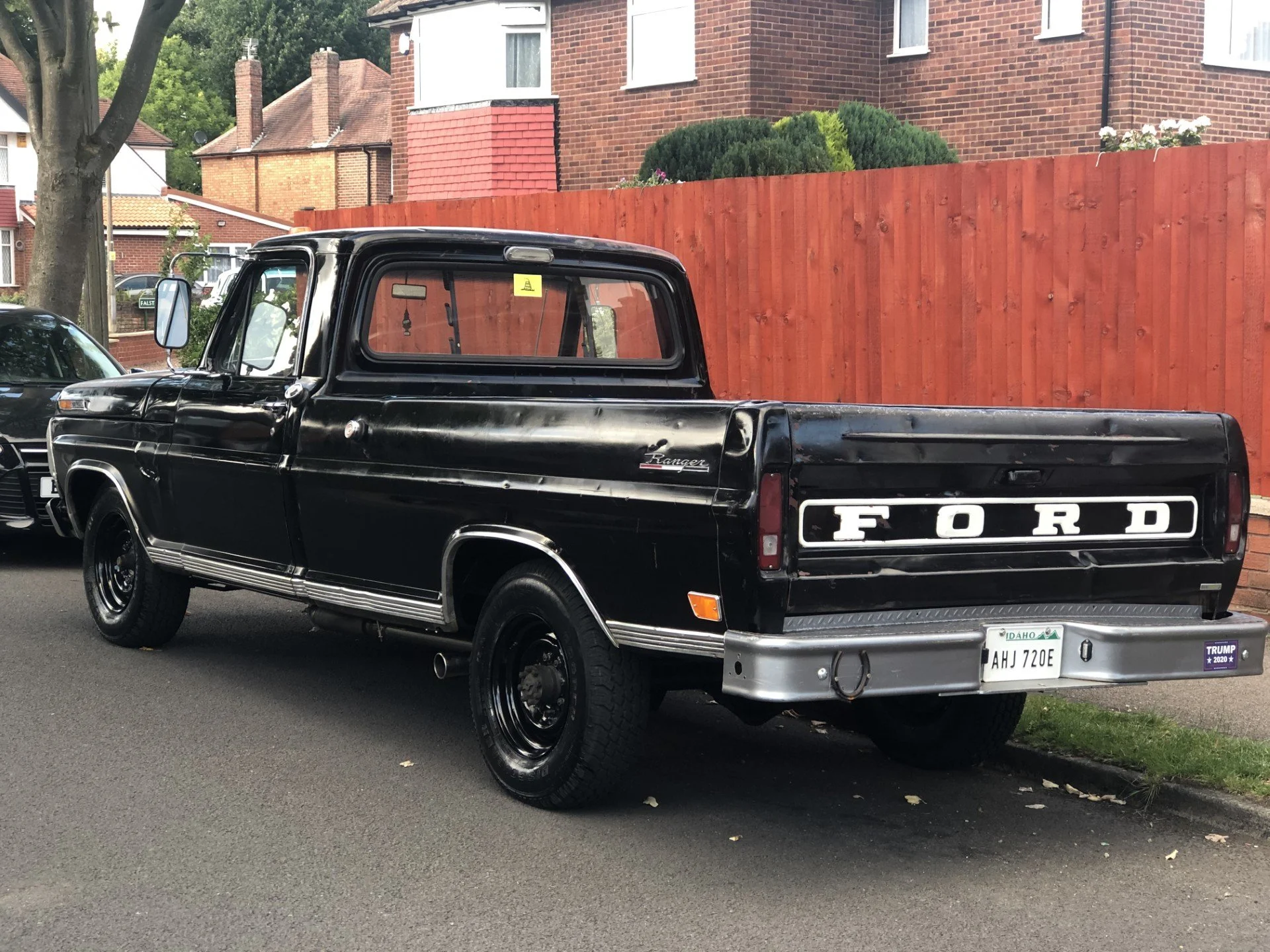 A black vintage Ford Ranger pickup truck parked on a residential street with a red wooden fence and brick houses in the background.