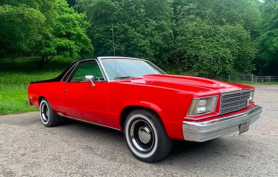 A vintage red convertible car parked on a gravel path surrounded by green trees.