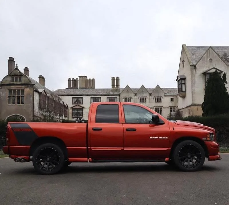 Red pickup truck parked in front of a large, old-style building with multiple roofs and chimneys on a cloudy day.
