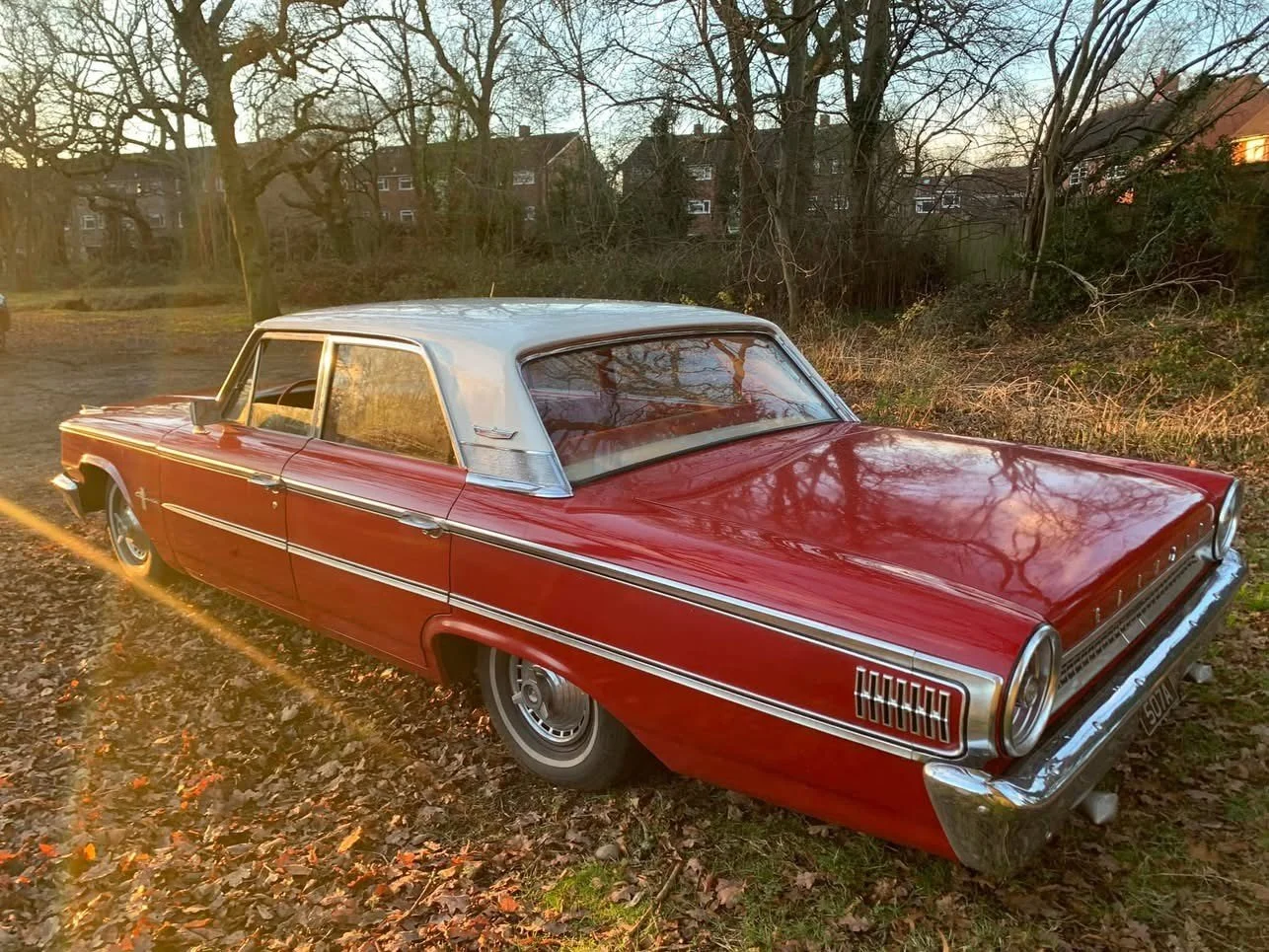 A vintage red and white two-tone car parked on a leaf-covered ground in a suburban area during sunset, with trees and houses in the background.