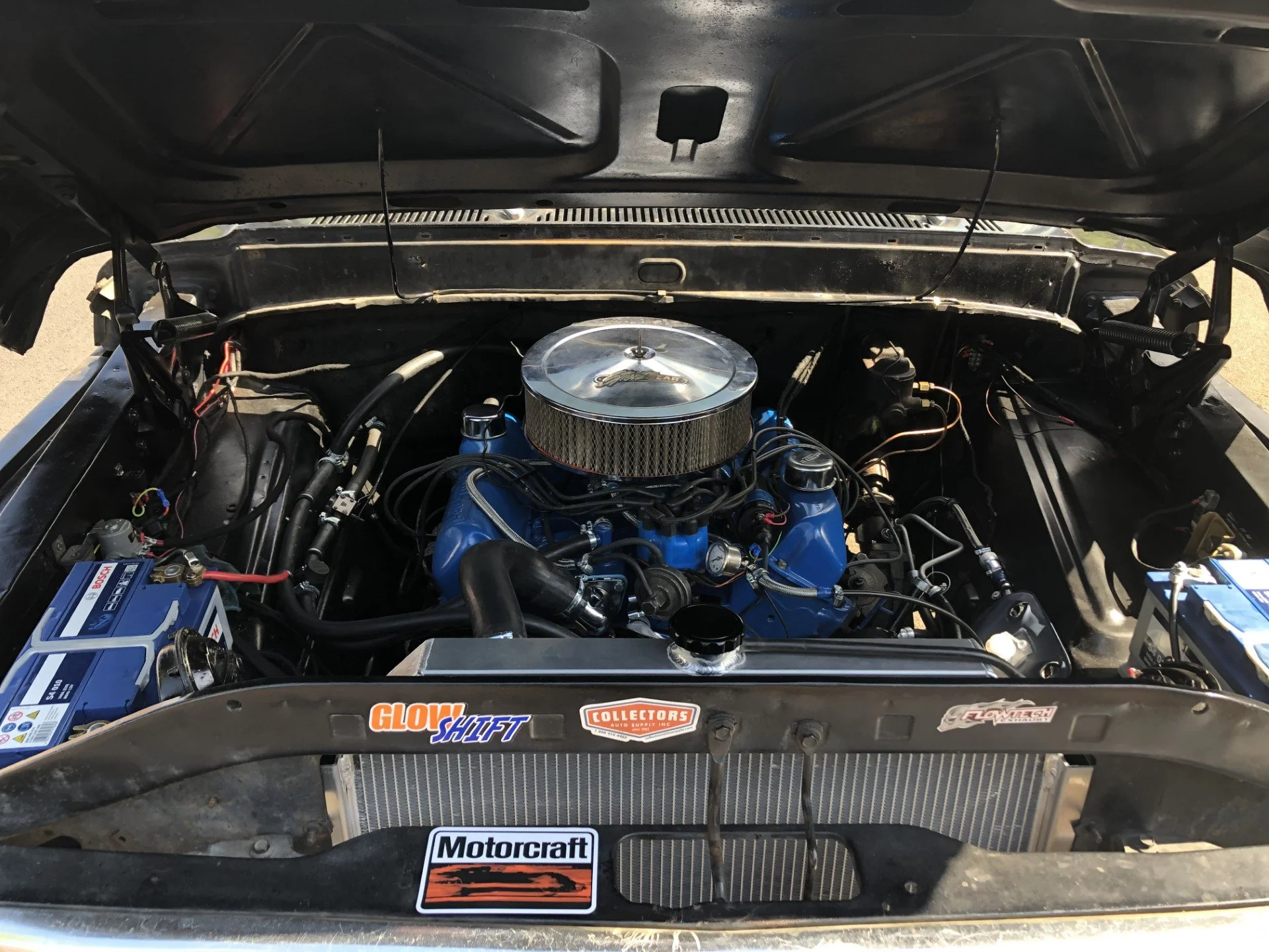 View of a car engine in the engine bay, featuring a chrome air filter cover, blue engine components, various wiring, and a radiator in the front. Stickers on the front include 'Glow Shift,' 'Collector's Auto Supply,' and 'Motorcraft.'