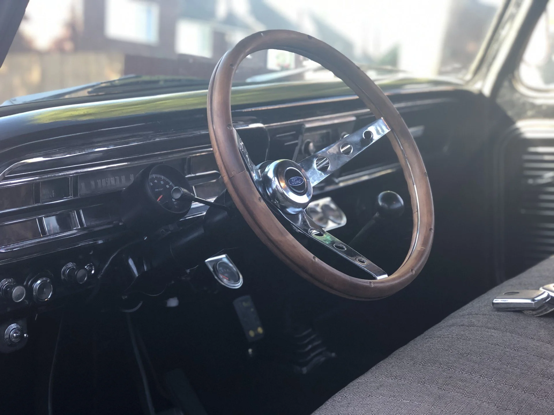 The interior of a vintage car, featuring a classic wooden steering wheel, a black dashboard with gauge, and various controls, with sunlight coming through the windows.