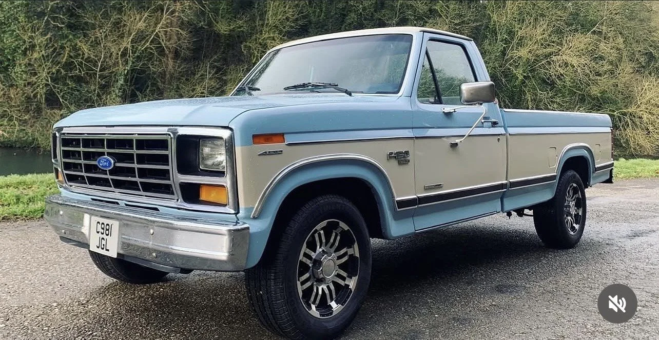 A vintage blue and beige Ford F-150 pickup truck parked on a gravel road with a grassy area and trees in the background.