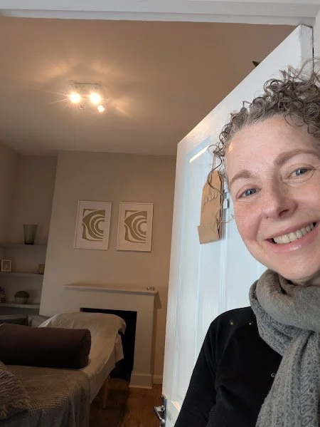 A woman with curly hair smiling inside a treatment room, standing near a partially open door at Grey Door Therapy. The background shows a treatment room with a beige couch, framed wall art, and some shelves with decorative items.