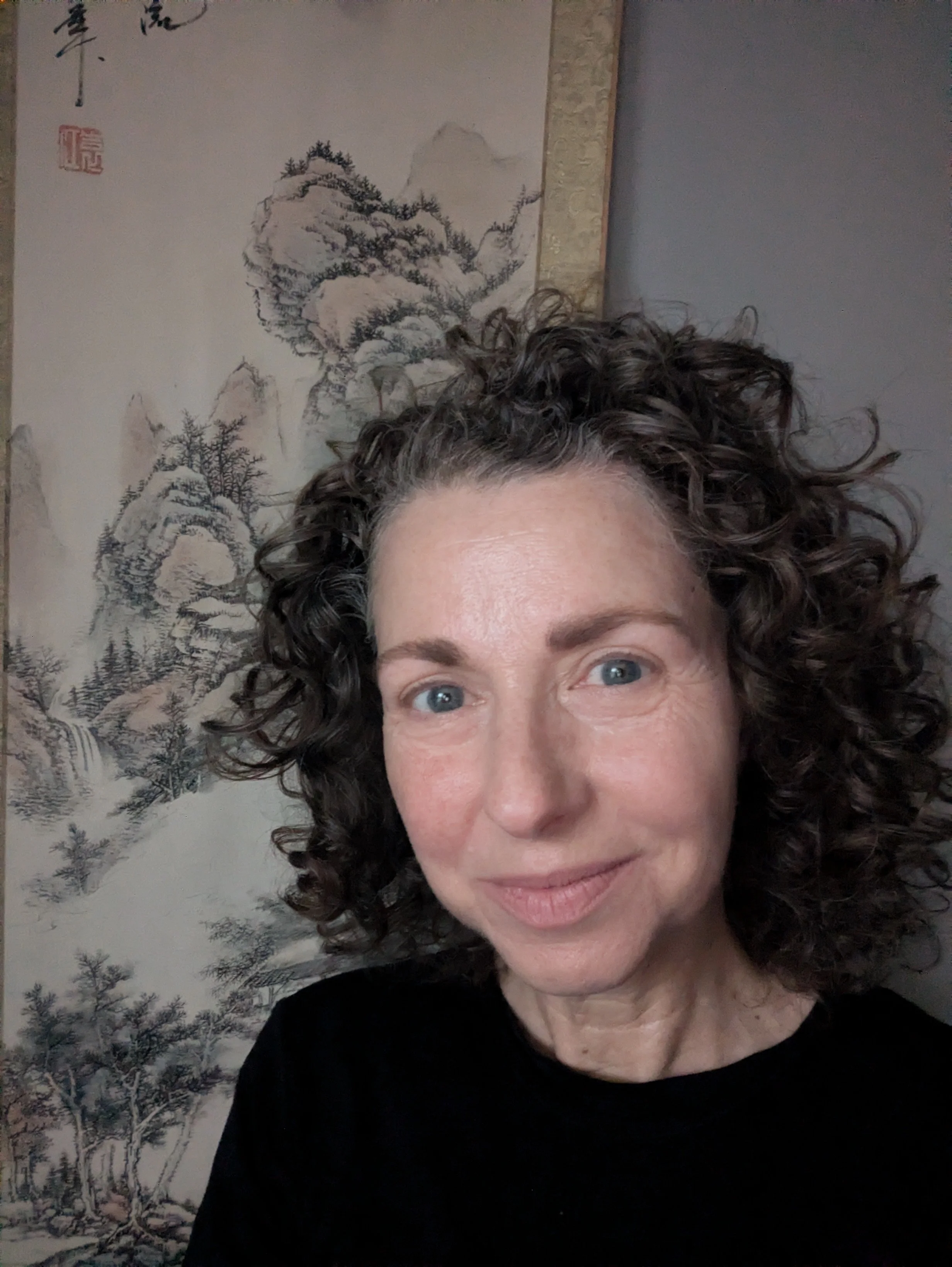 Close-up of a woman with curly hair and blue eyes, with a mountainous landscape painting in the background.