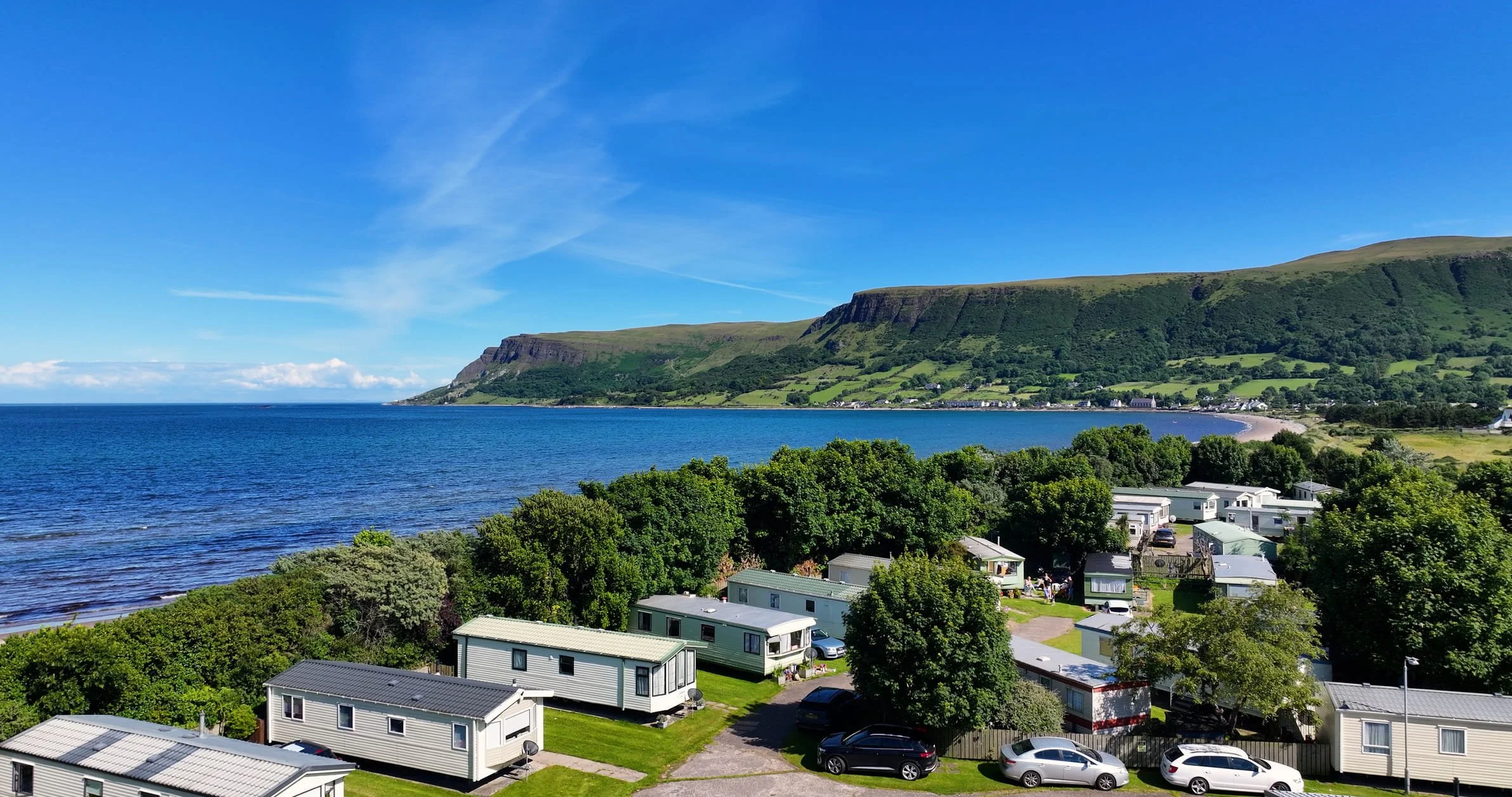 A scenic coastal view of a bay with green hills and cliffs in the background, a blue sky with some clouds, and a row of white residential buildings with cars parked in front.