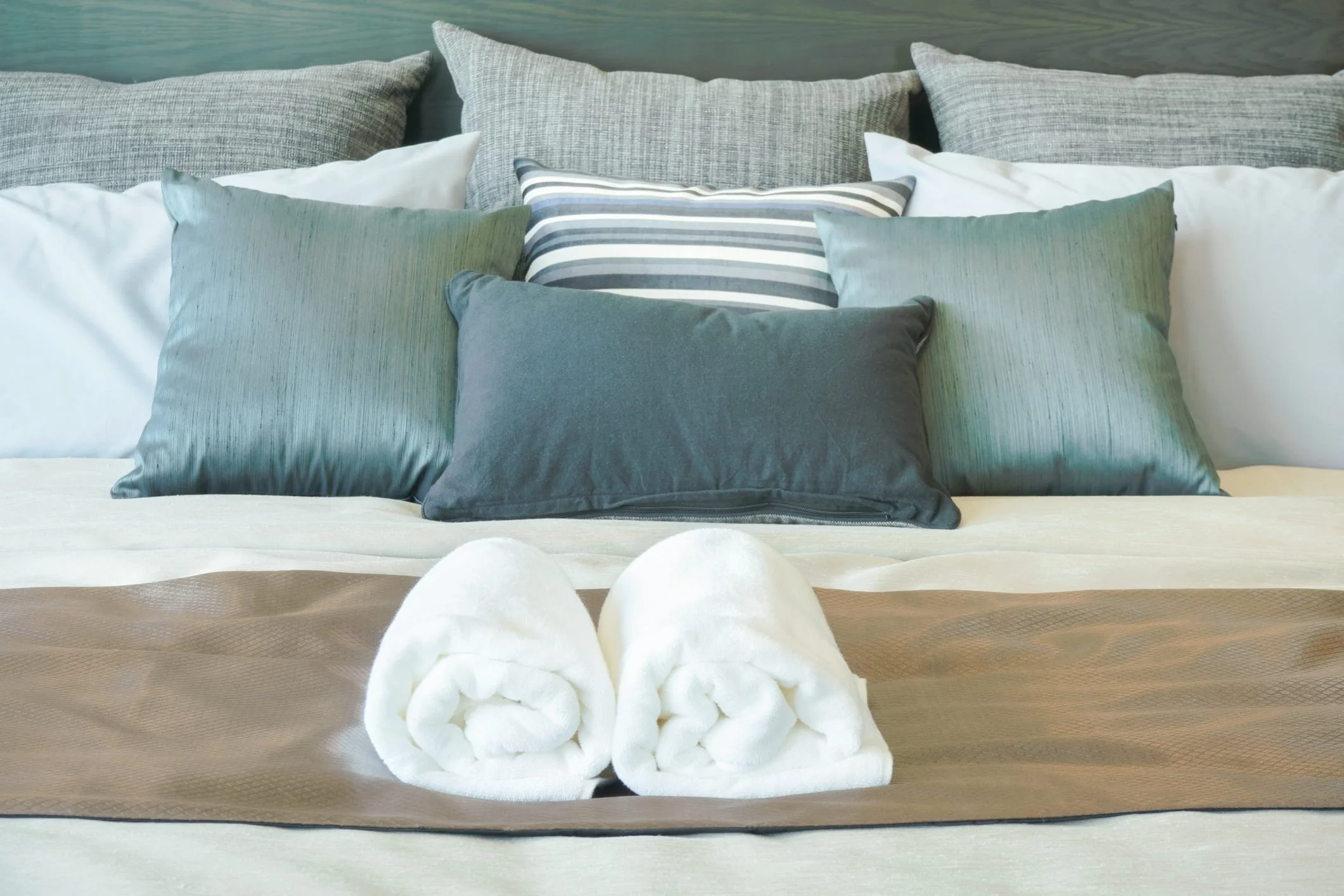 A neatly folded towel sculpture resembling a bouquet of flowers, placed on a bed with multiple pillows in shades of blue, gray, and white.