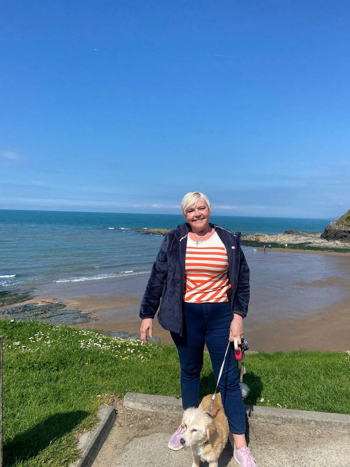A woman with short white hair smiling and standing on a grassy area by the beach, holding a leash with a small golden dog with a pink bandana, with the ocean and rocky shoreline in the background under a clear blue sky.