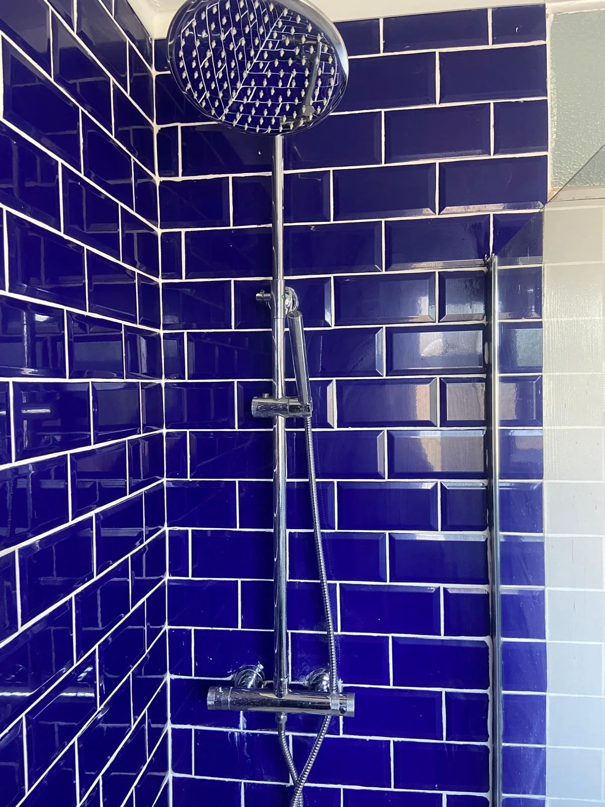 A modern shower with dark blue subway tiles on the walls, a round showerhead, a handheld shower attached to a vertical bar, and a glass door edge.