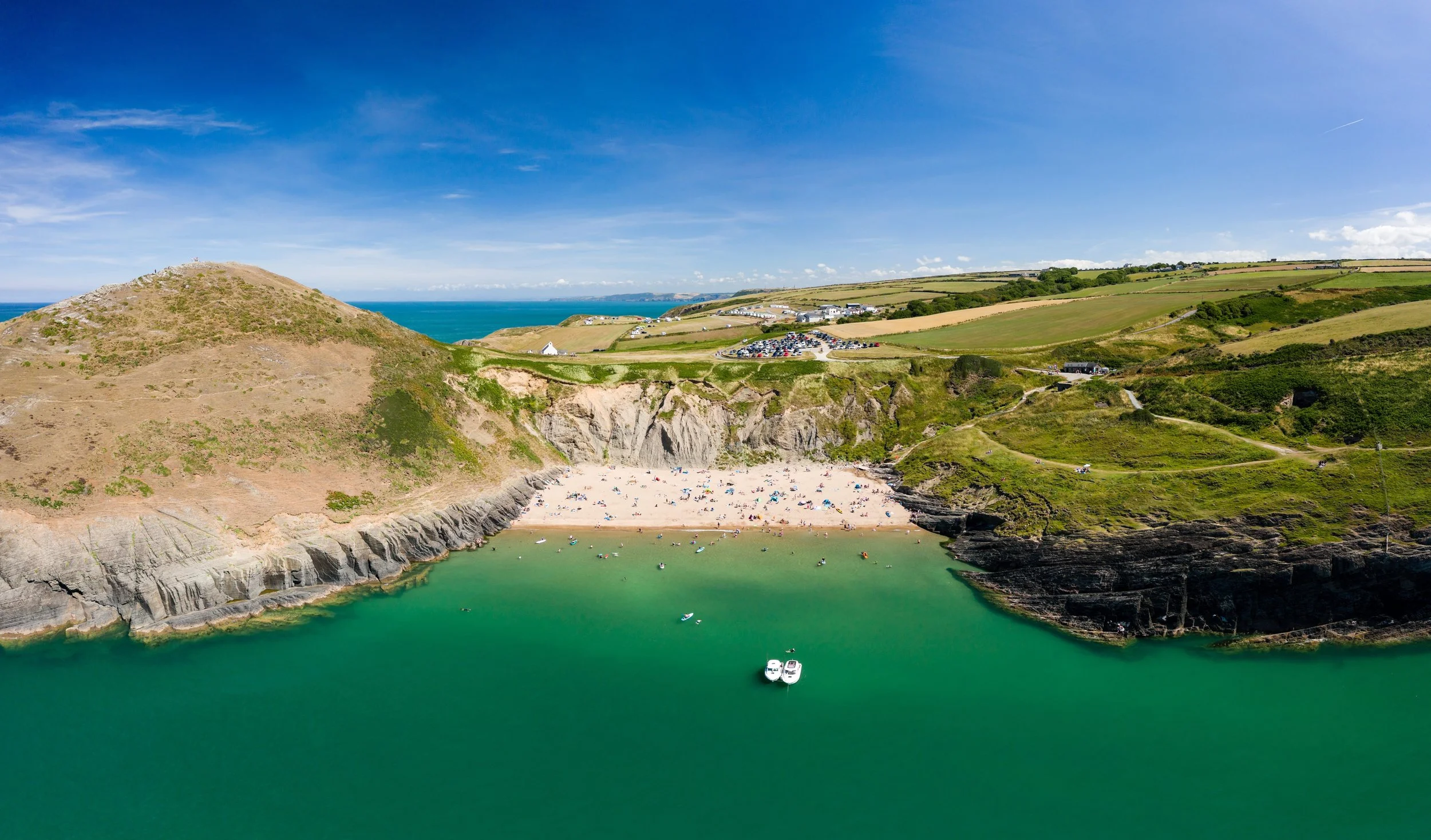 Scenic coastal landscape with a beach, cliffs, green hills, and a parking lot under a blue sky.