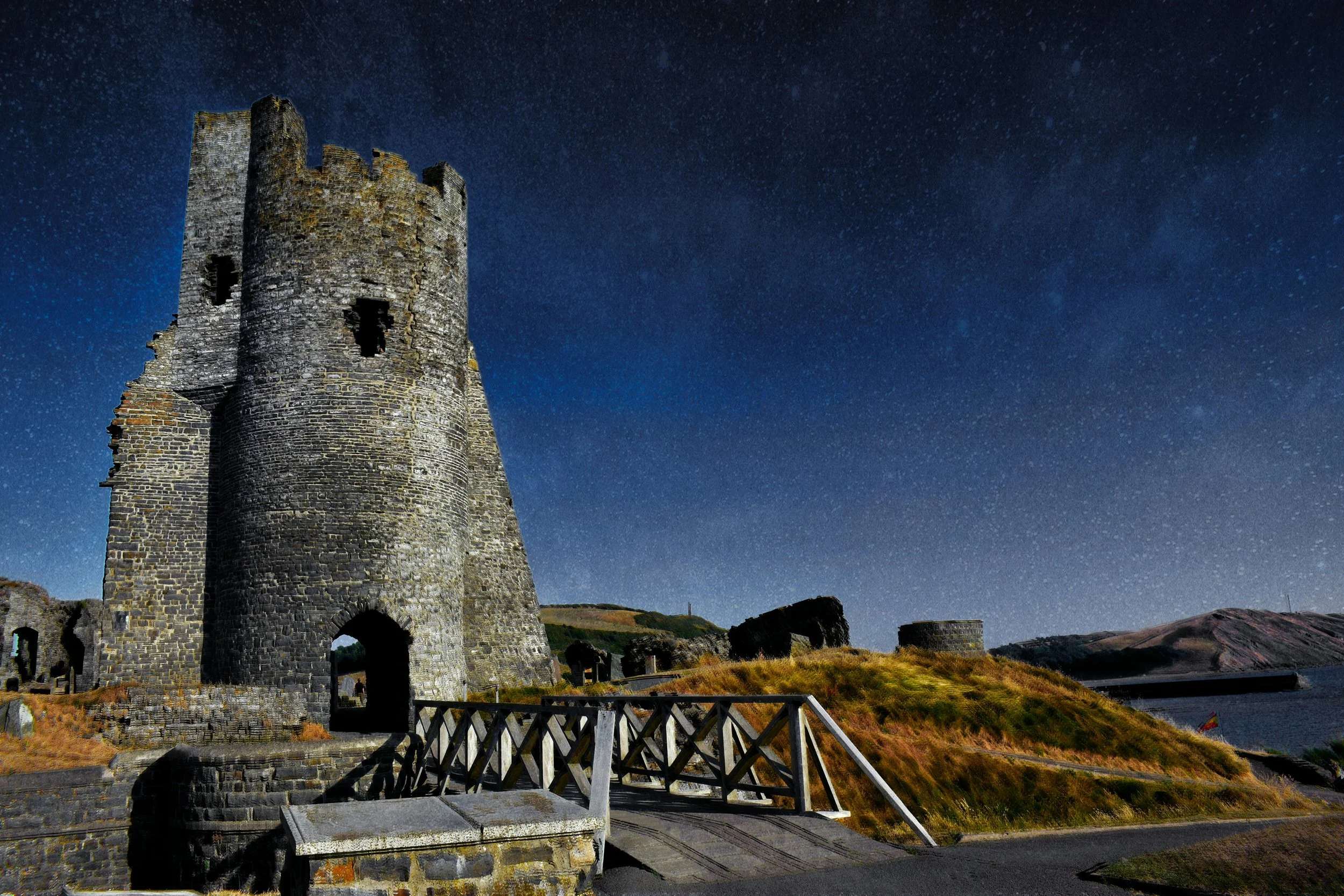 Ancient stone castle tower under a starry night sky, with surrounding ruins and a wooden fence in the foreground.