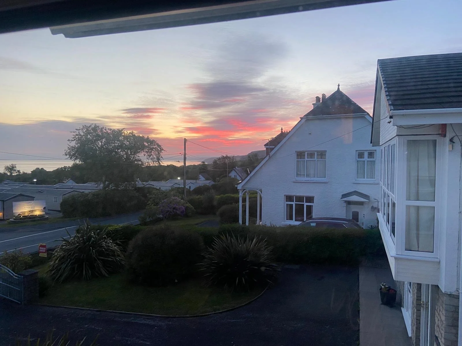 A view of a residential neighborhood at sunset with a large tree, white house with bay windows, and a sky filled with colorful clouds.