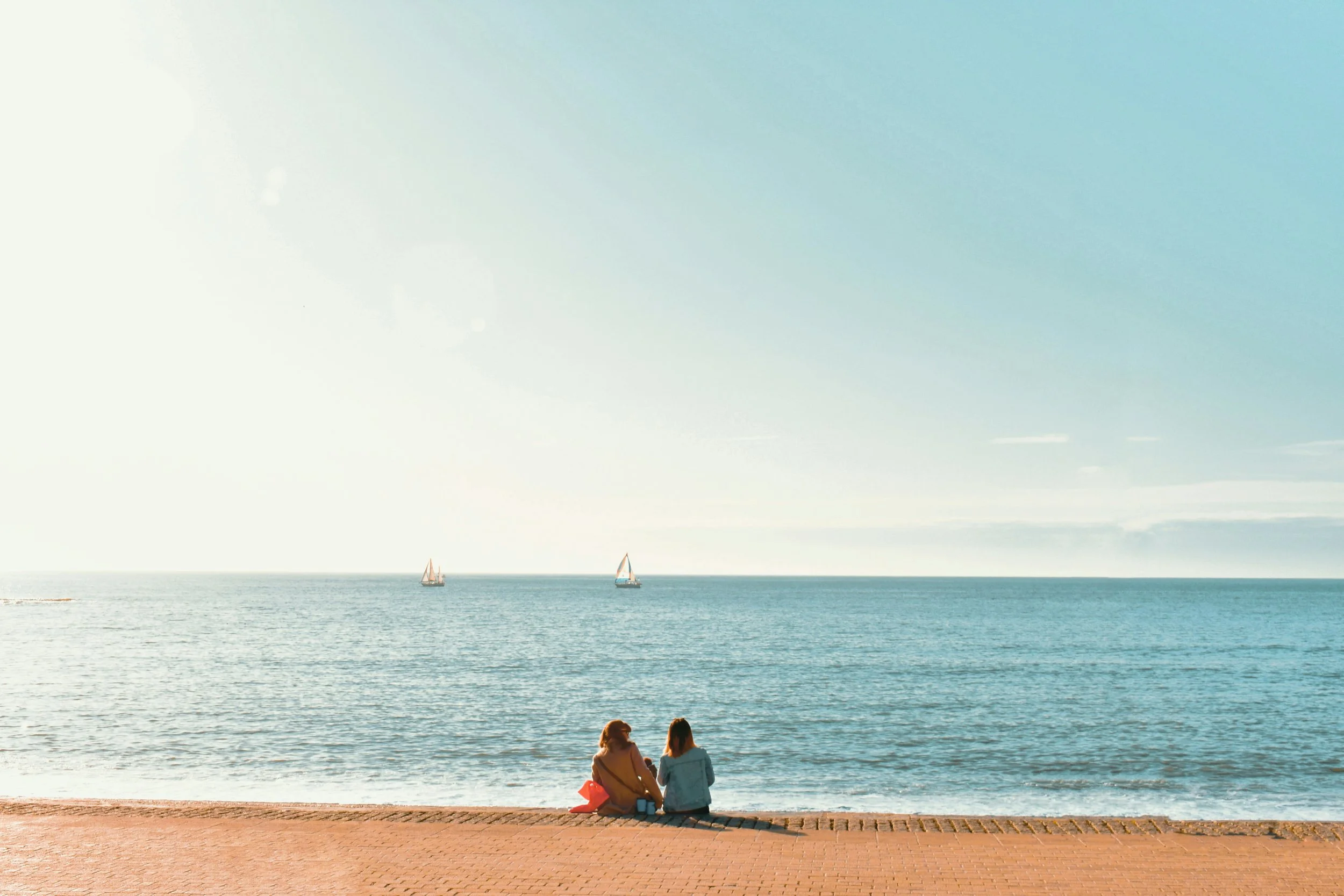 Two women sitting on a beach with sailboats on the water and a partly cloudy sky in the background.