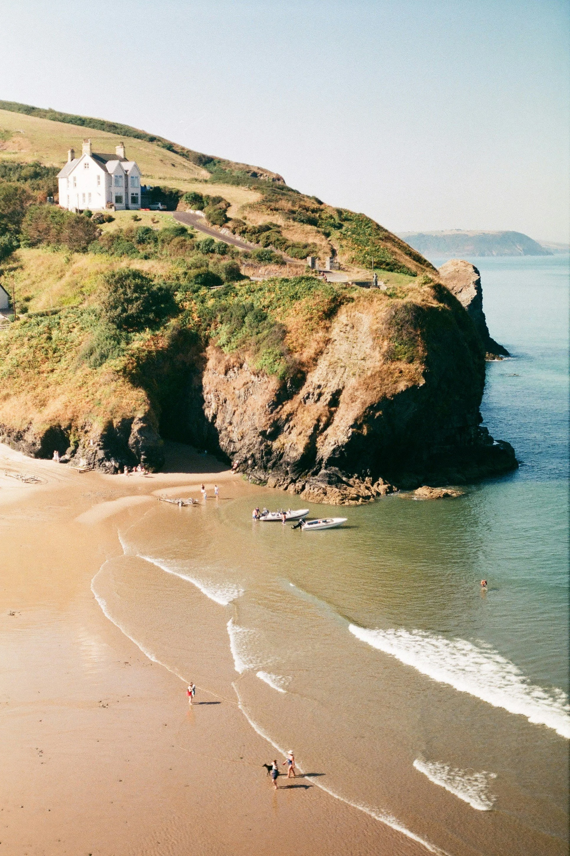 People walking along sandy beach with small boats anchored in shallow water, cliffs in the background under a clear sky.