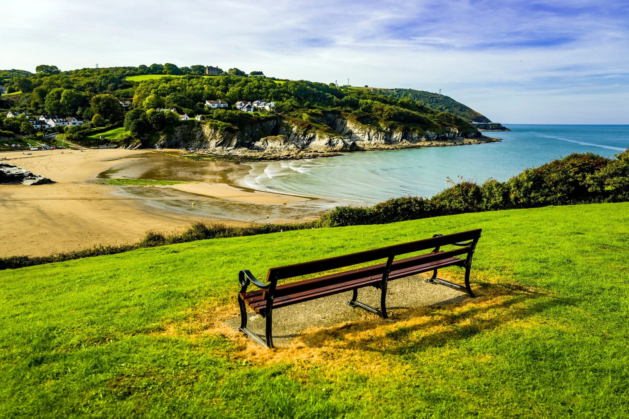 A grassy park with a bench, overlooking a beach and rocky coastline with houses and green hills in the background.