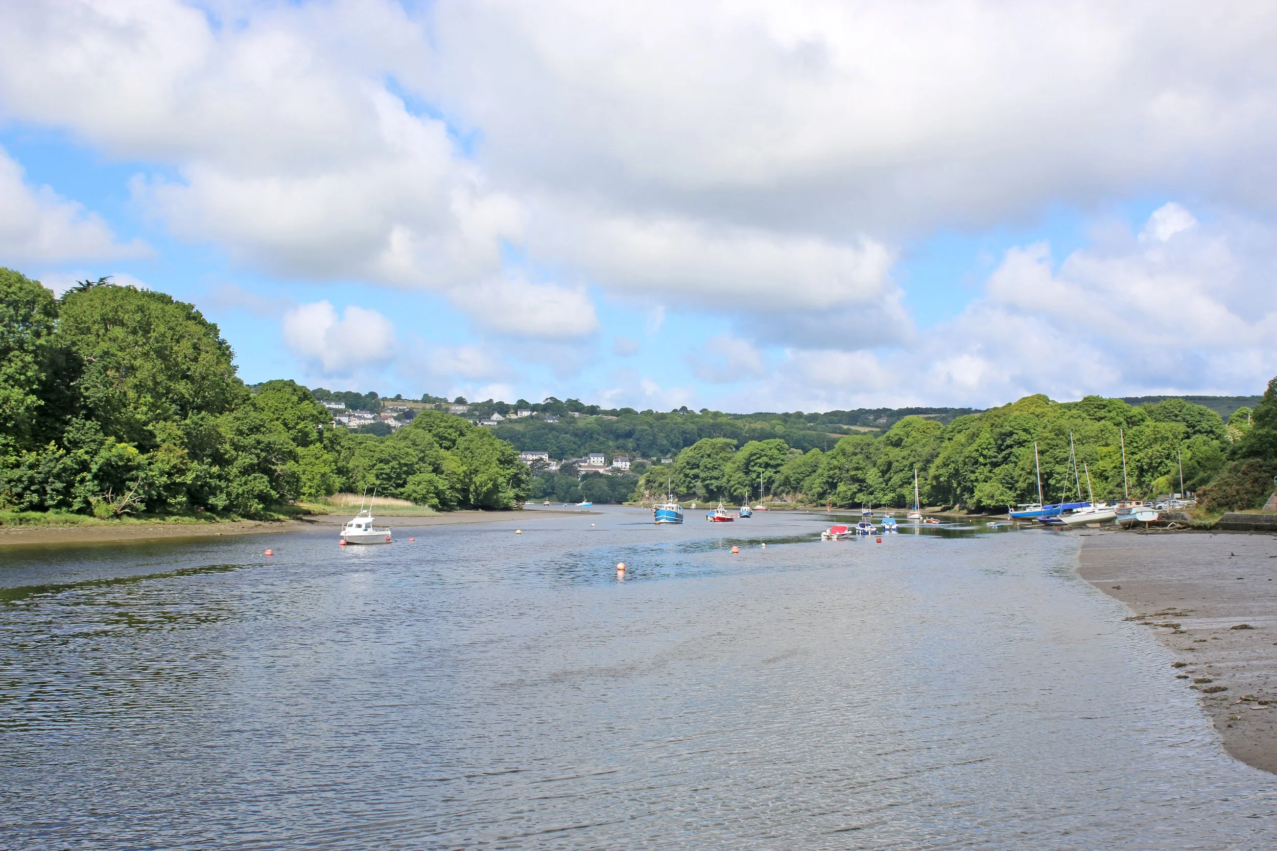 A scenic view of a river with small boats docked along the shoreline surrounded by lush green trees under a partly cloudy sky.