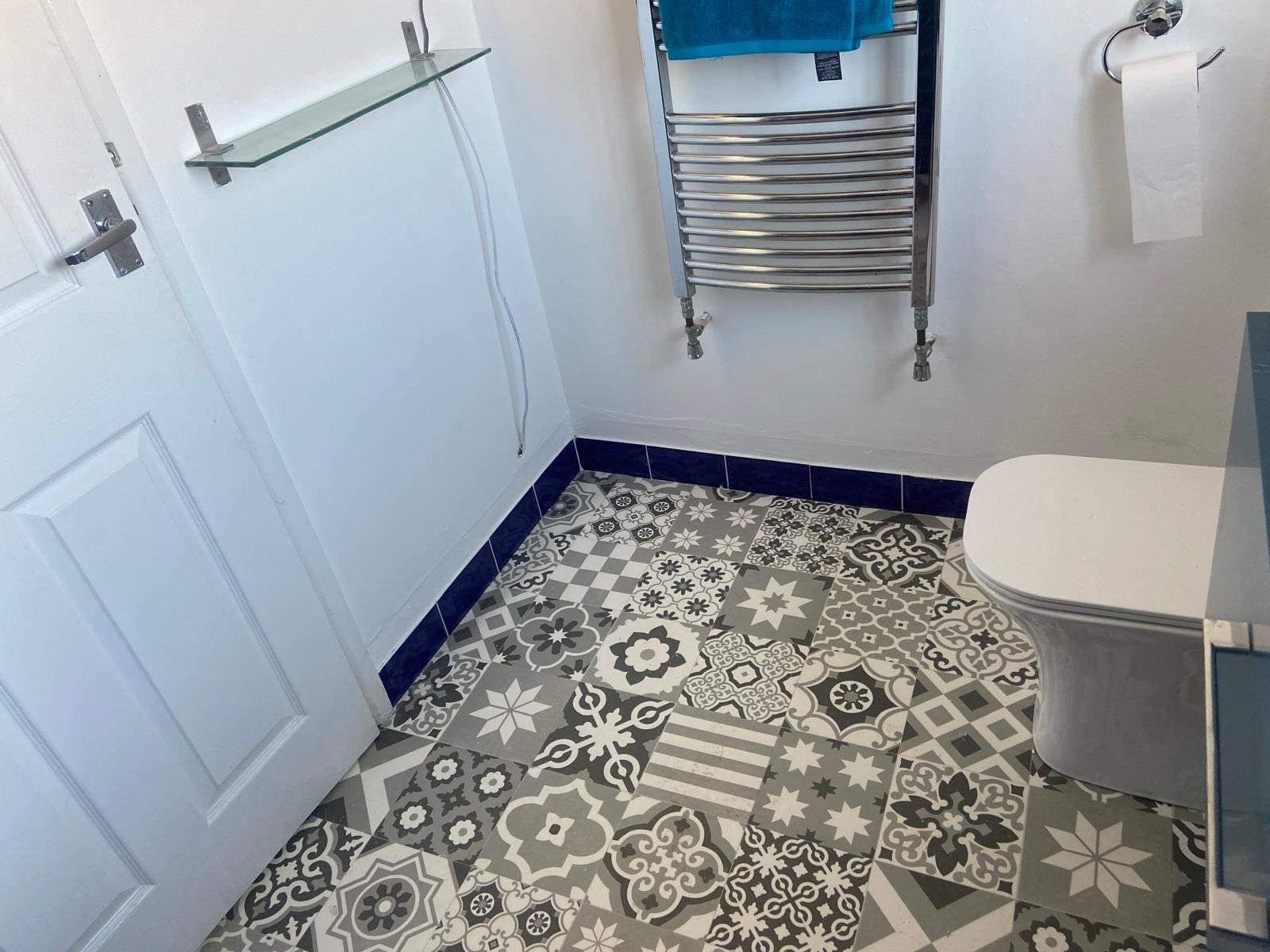 A bathroom with black and white patterned floor tiles, a white toilet, a wall-mounted towel rack with a blue towel, a glass shelf, and a toilet paper holder with a roll of toilet paper.