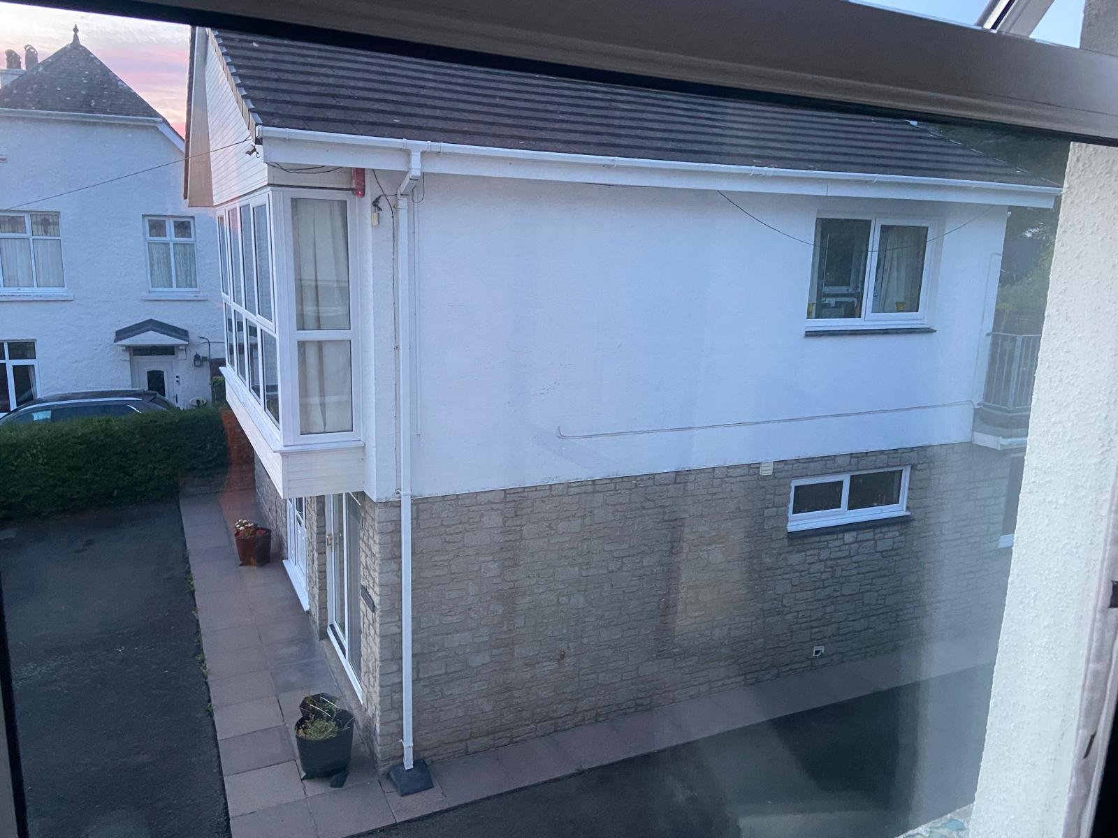 View of a three-story residential building seen through a window with a glass door in the foreground. The building has a brick lower level and a white upper level with several windows, including a small stained glass window at the top. The house is a