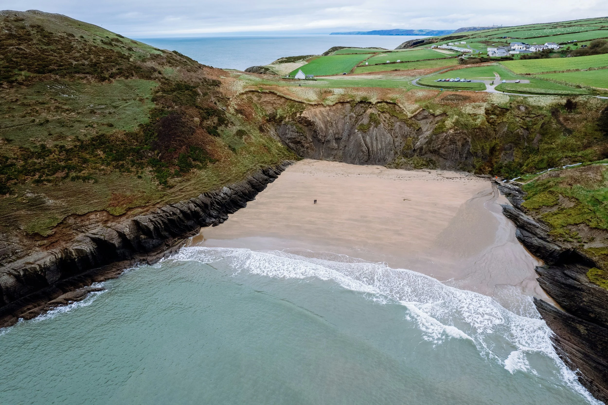 Cliffside beach with waves crashing against rocks, green hills and farmland in the background, and a view of the ocean and sky.