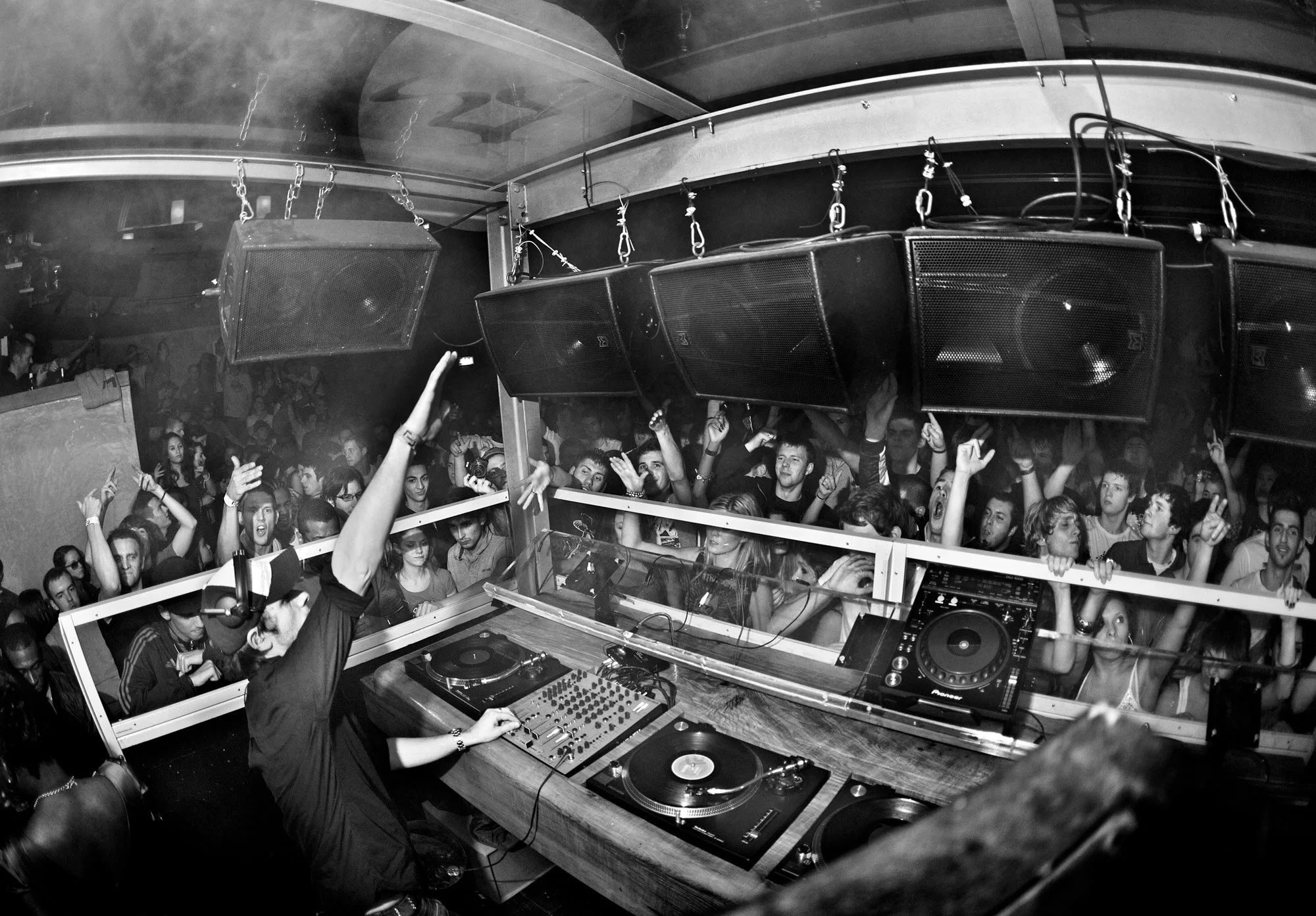 DJ Andy C performing at a Matter nightclub, London with a large crowd dancing and raising their hands; the DJ is using turntables and mixers, and there are large speakers hanging from the ceiling.