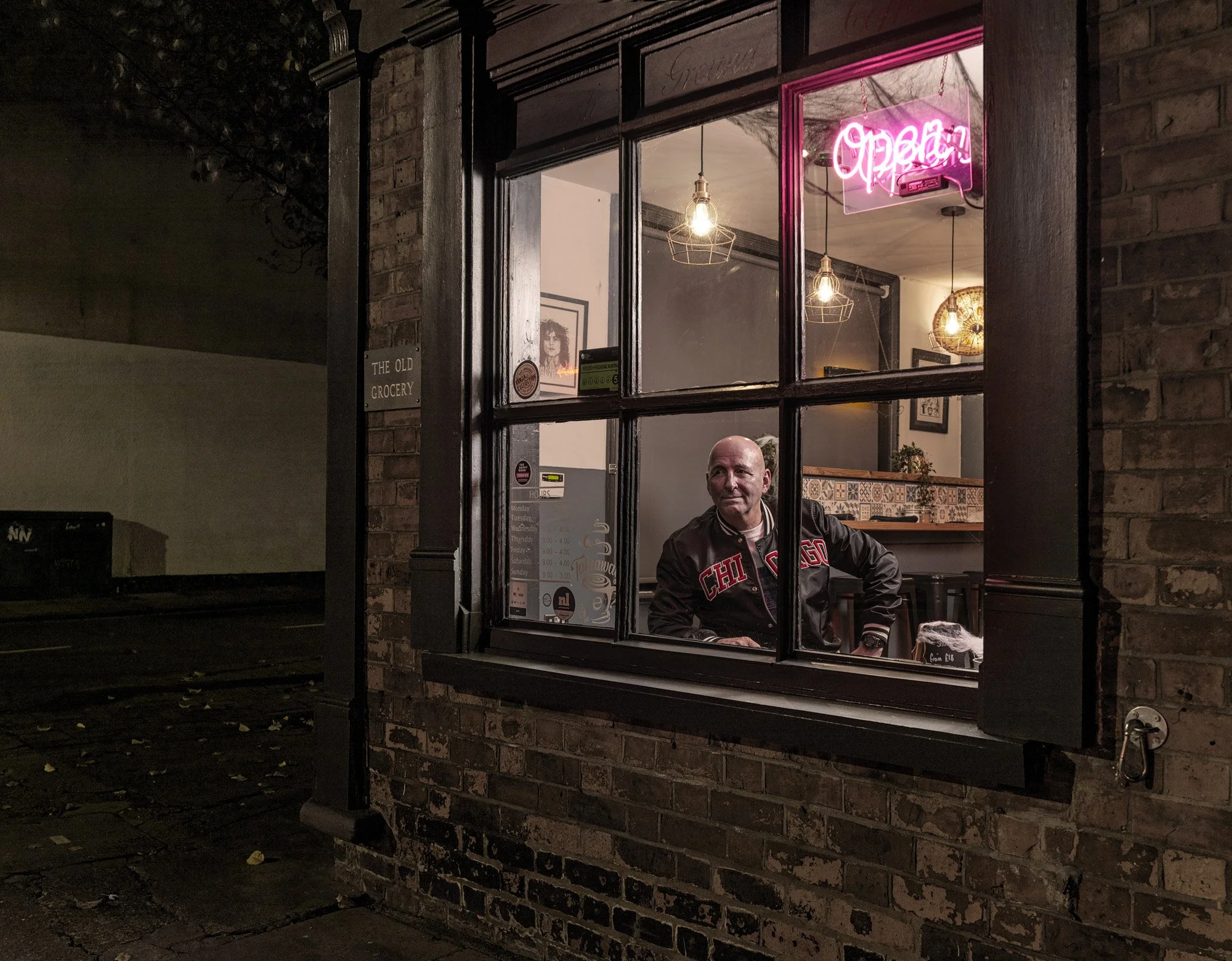 Nighttime view of a small eatery or cafe with a brick exterior and a large window. Inside, a man wearing a jacket with "CHICAGO" on it is smiling. The interior has hanging pendant lights, framed artwork, and a festive wall decoration. A bright pink "OPEN" neon sign is visible in the window.