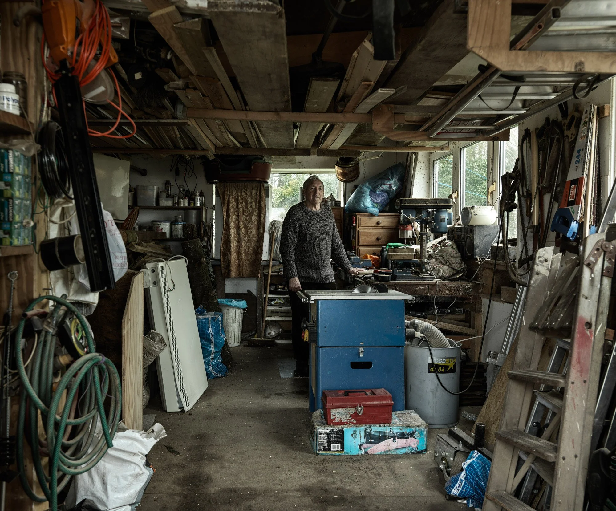 A person standing in a cluttered workshop or garage with various tools, equipment, and supplies, including a drill press, shelves filled with cans and bottles, and workbenches, surrounded by wood and miscellaneous items.