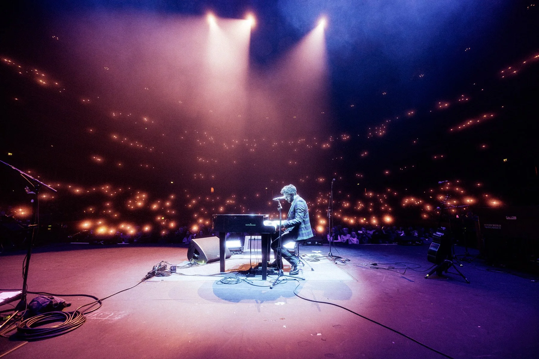 Billy Lockett playing a keyboard on stage, under purple and blue spotlights, at the Royal Albert Hall, filled with glowing lights and an audience.