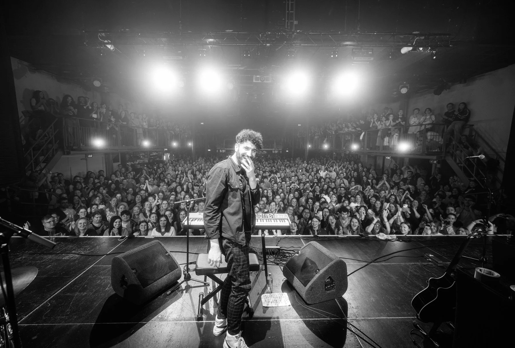 Billy Lockett  on stage during a concert in Amsterdam, smiling and covering part of their face with one hand, with a large crowd of fans in the background, cheering and clapping, in a concert hall with bright overhead lights.